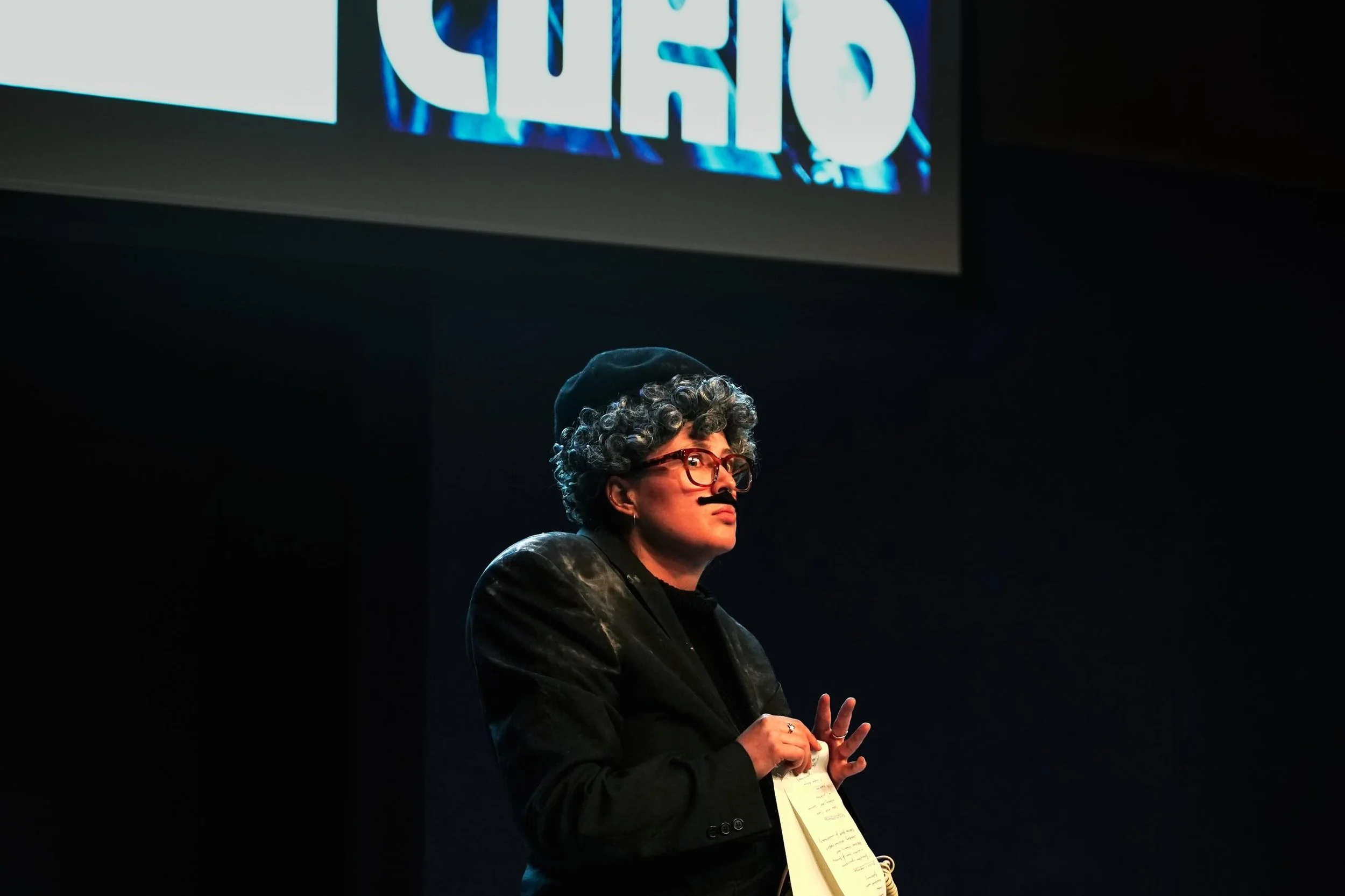 Performer Izzy Stott is on stage as alter ego Gunter, dressed in a black beret and fake moustache