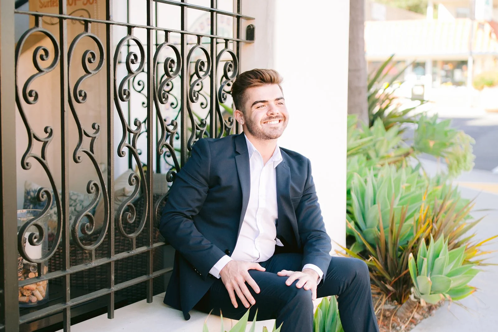 Senior student sitting on edge of building in suit looking off into the distance surrounded by  plants taken by GunnShot Photography