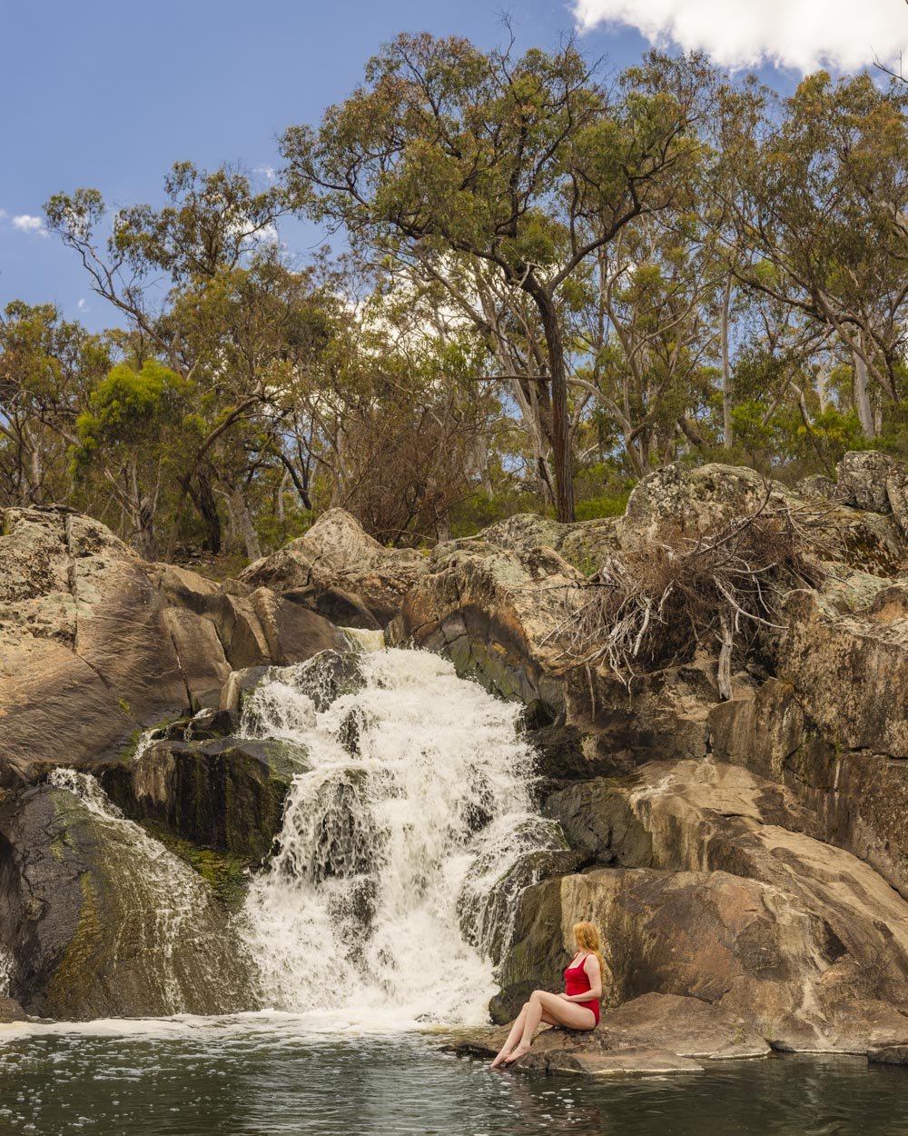 Lady in a red swimsuit sitting on rocks by a pool at the base of the waterfall.