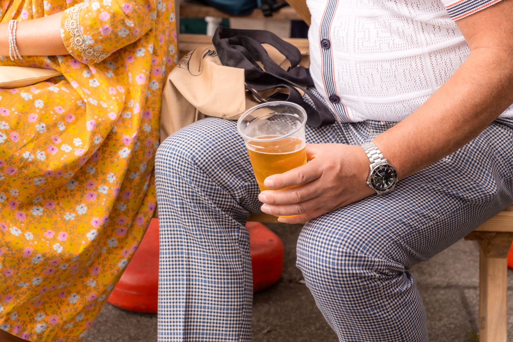 A man sitting on a wooden bench holding a plastic cup of beer. He is wearing a white patterned shirt, checkered pants, a silver watch, and a silver bracelet. Part of a woman in a yellow floral dress is visible next to him.