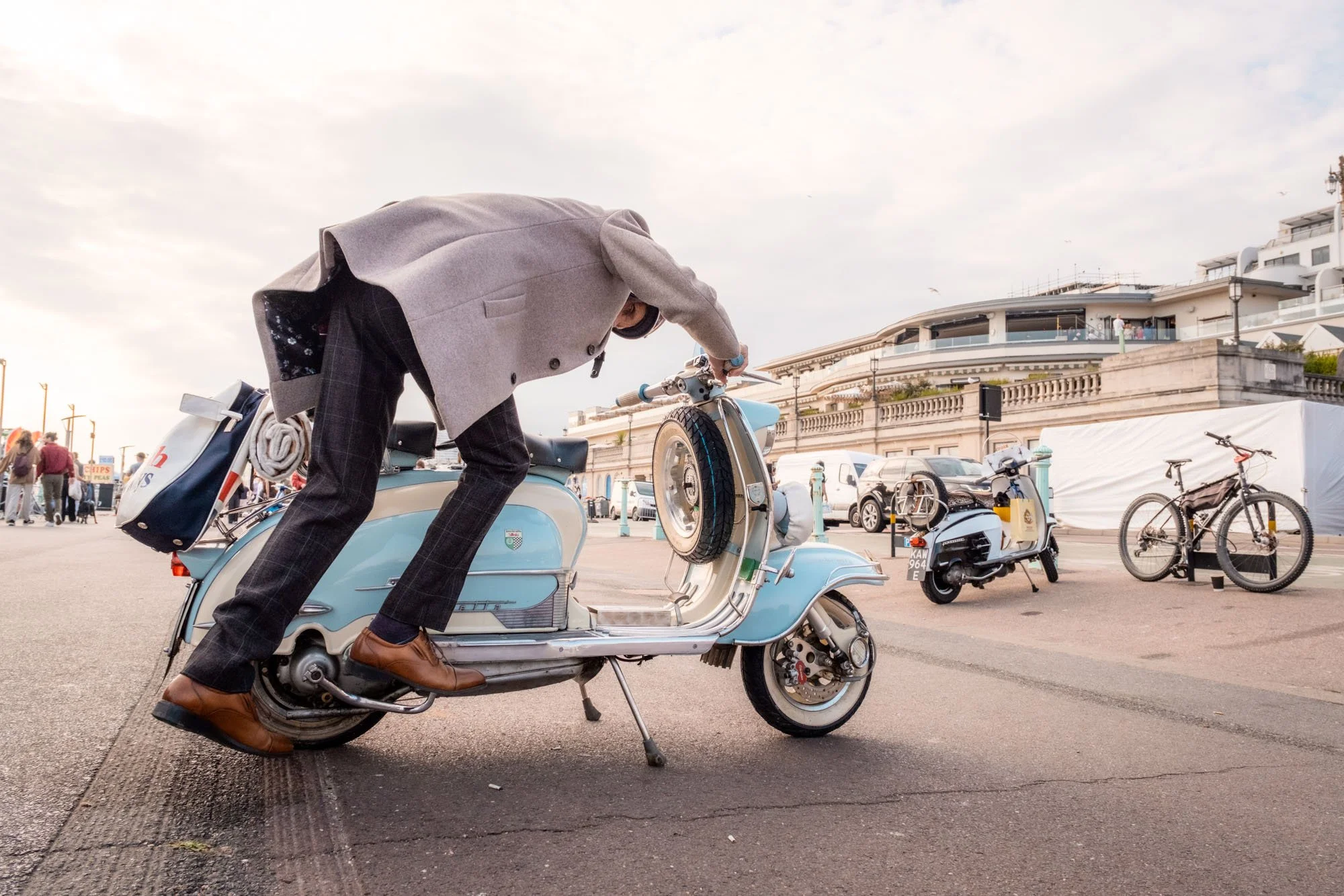 A person in a gray coat and brown shoes standing on a blue vintage scooter, adjusting the handlebar, with others and buildings in the background.