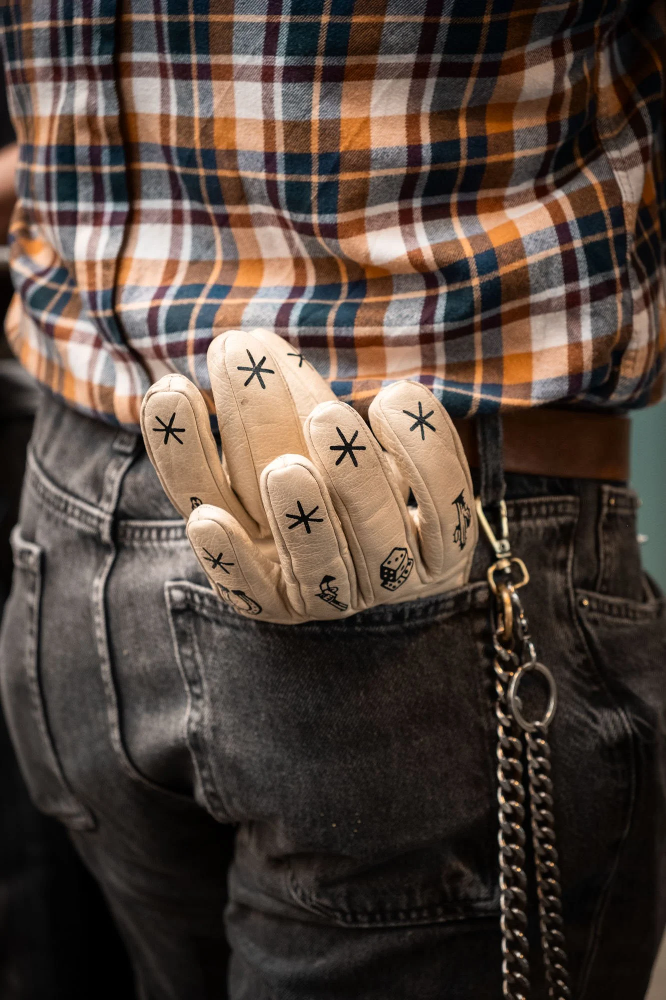 A person wearing black jeans and a plaid shirt has a pair of beige gloves with black symbols in their back pocket, with a silver chain attached to the jeans.