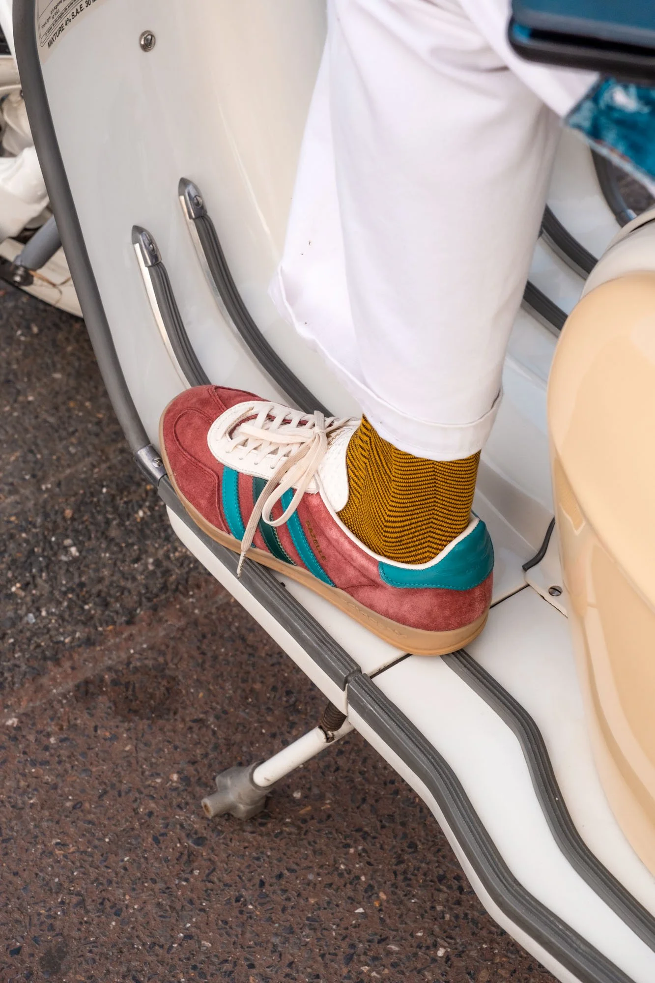 A colorful sneaker with red, teal, and beige accents worn over mustard yellow and black striped socks, on a motorcycle footrest.