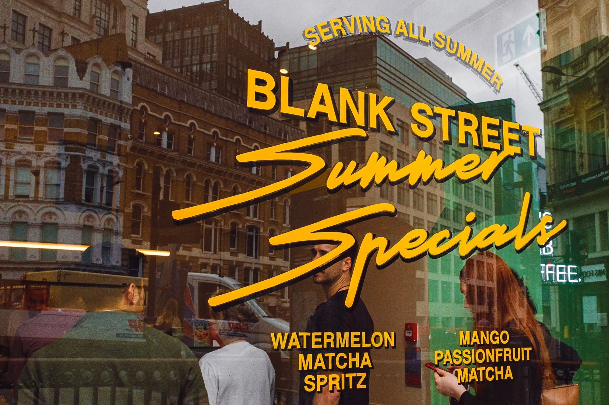 Reflection of a street scene on a glass window, with yellow text advertising summer specials for fruit-flavored drinks, including watermelon matcha spritz and mango passionfruit matcha, with city buildings and pedestrians visible through the glass.