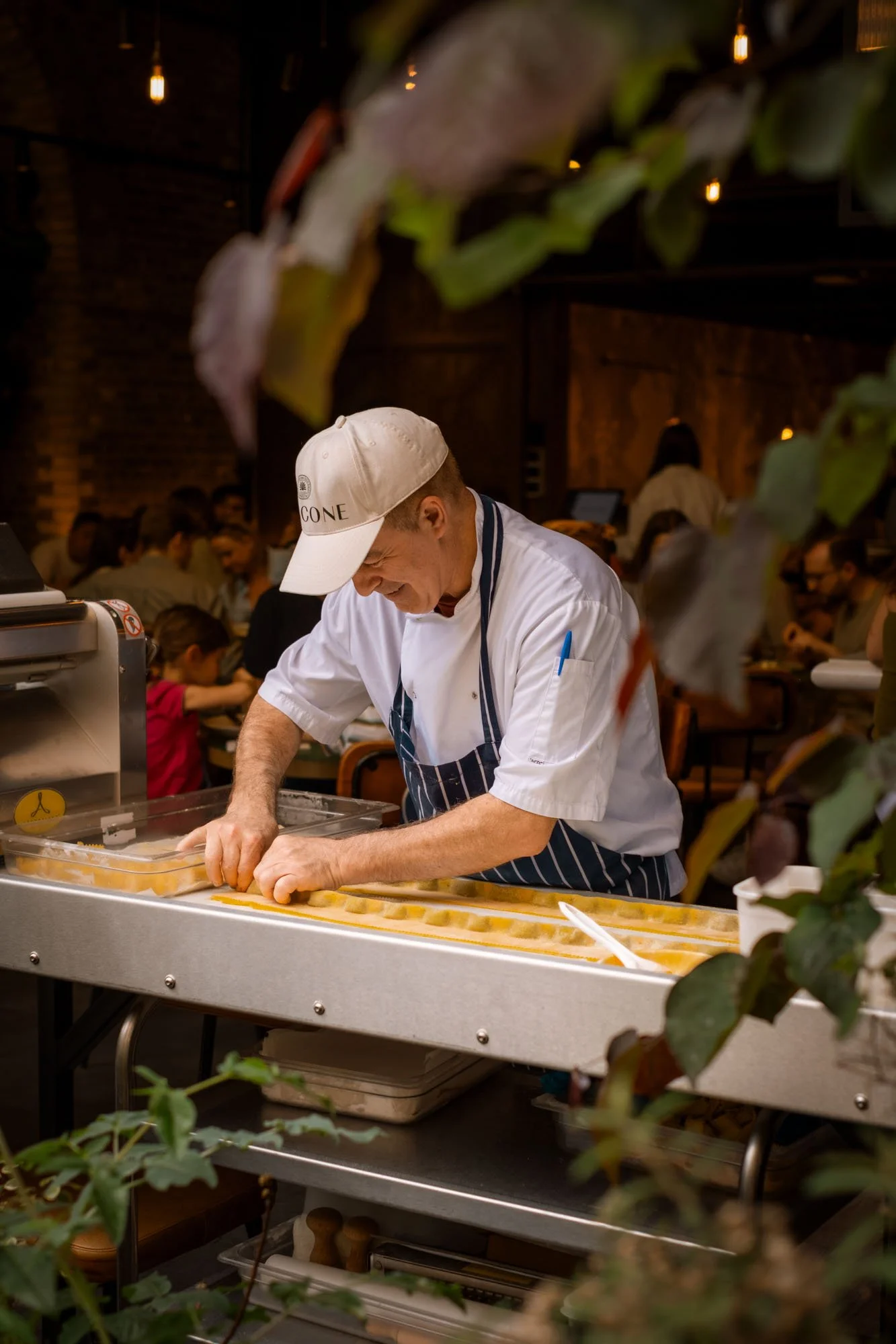 A chef wearing a white cap and striped apron makes pasta in a restaurant kitchen, surrounded by an environment filled with diners seated at tables.