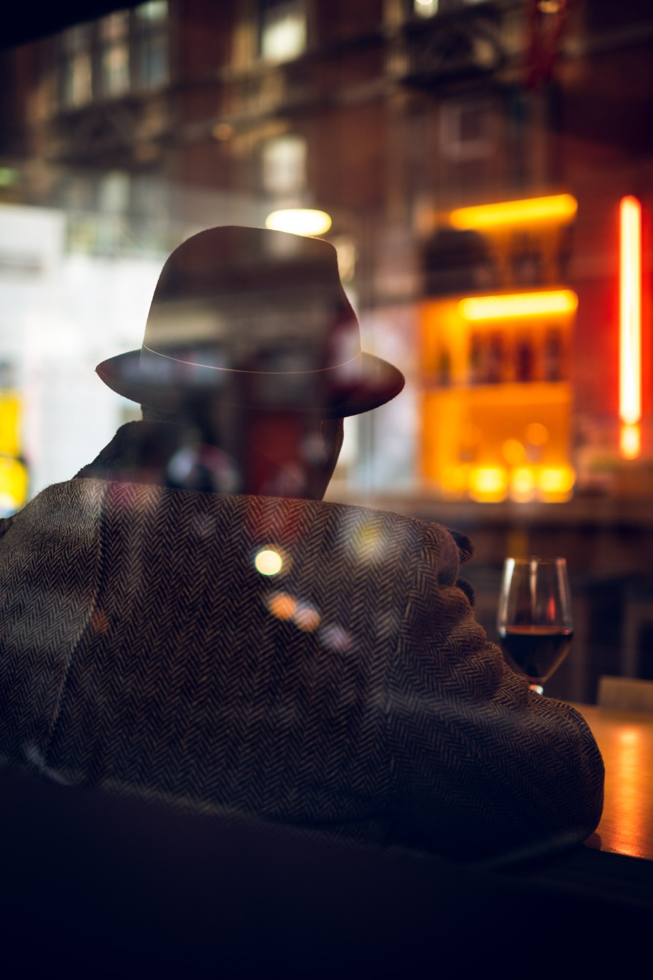 A person wearing a hat and a herringbone patterned coat sitting at a bar or restaurant, with a glass of red wine in front of them, depicted through a window or reflective surface with colorful lights and blurred city background.