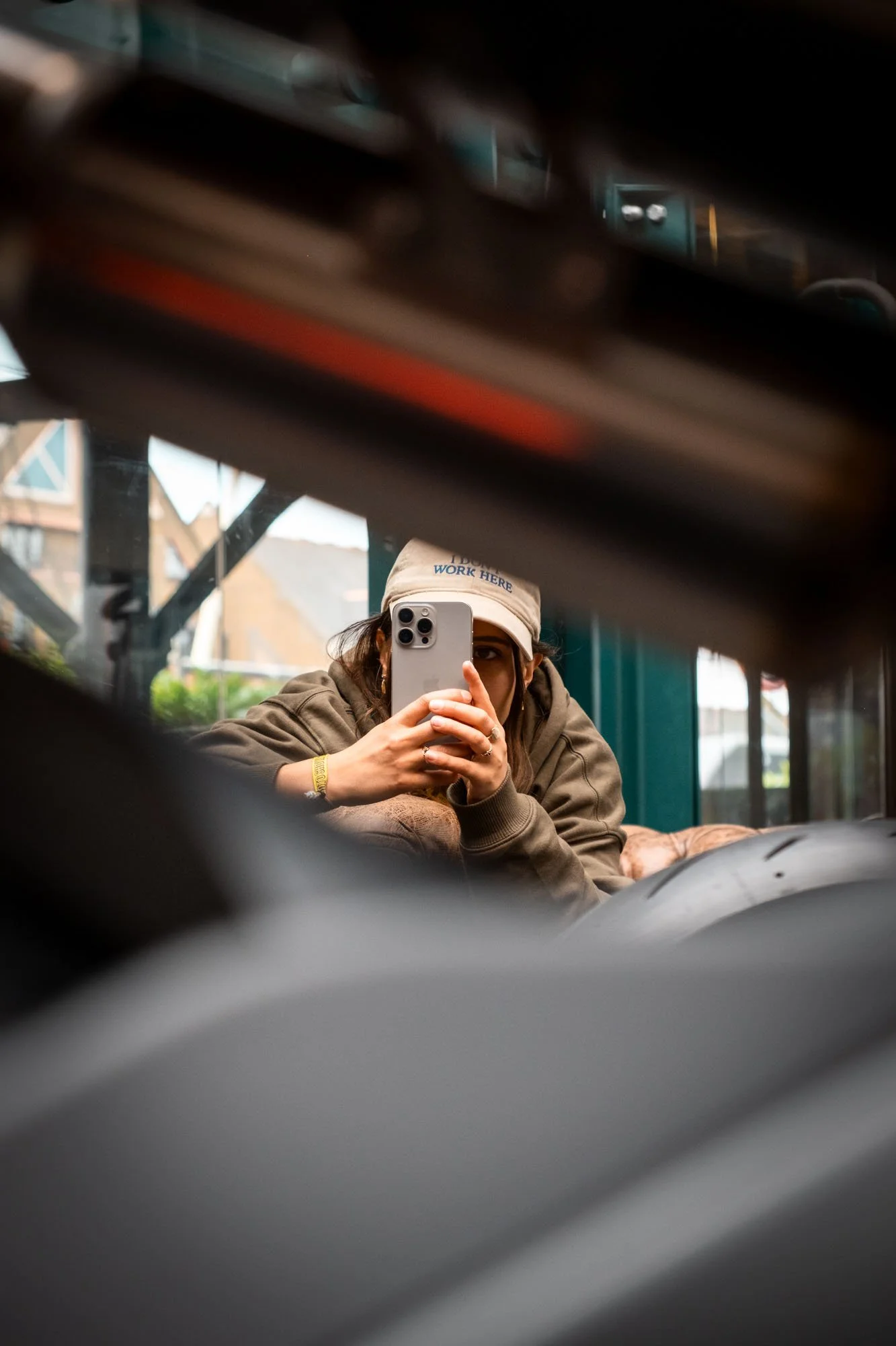 A person taking a selfie with a smartphone, wearing a beige cap and a hoodie, seen through a reflective or transparent surface with out-of-focus objects in the foreground.