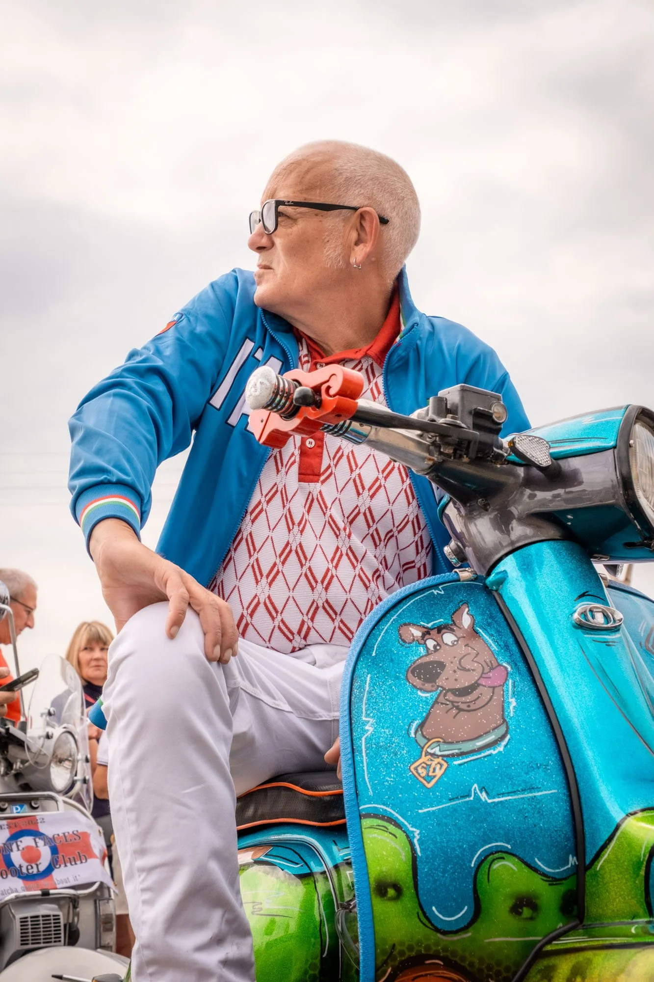 A person with light skin, glasses, and a shaved head sitting on a blue scooter decorated with a cartoon dog sticker, wearing a blue jacket and patterned shirt, looking to the side at an outdoor event with other people in the background.