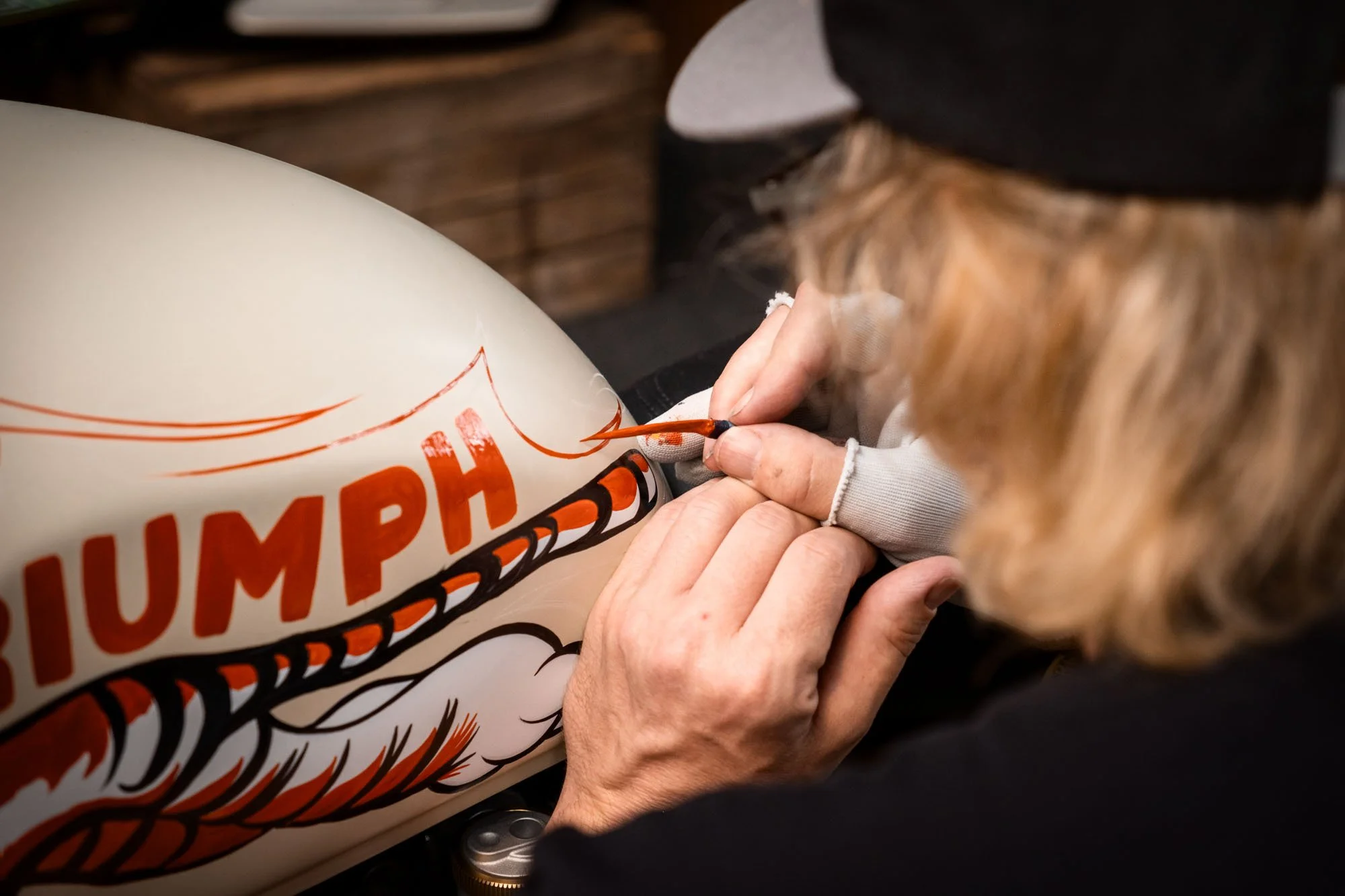 A person wearing a black cap and light-colored gloves is painting orange and black lettering on a white motorcycle fuel tank. The word 'TRIUMPH' is partially visible.
