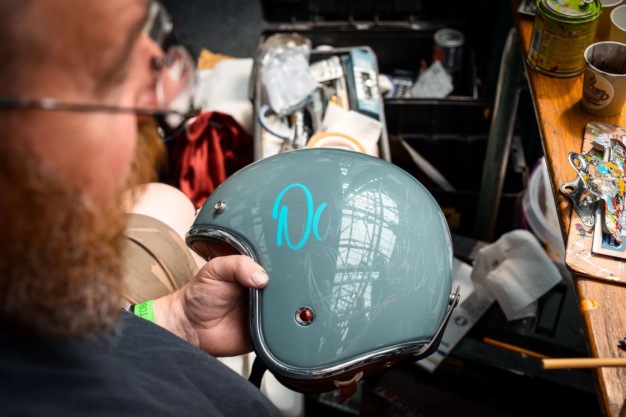 A man holding a gray motorcycle helmet with blue writing on it, in a workshop surrounded by paints, tools, and other supplies.