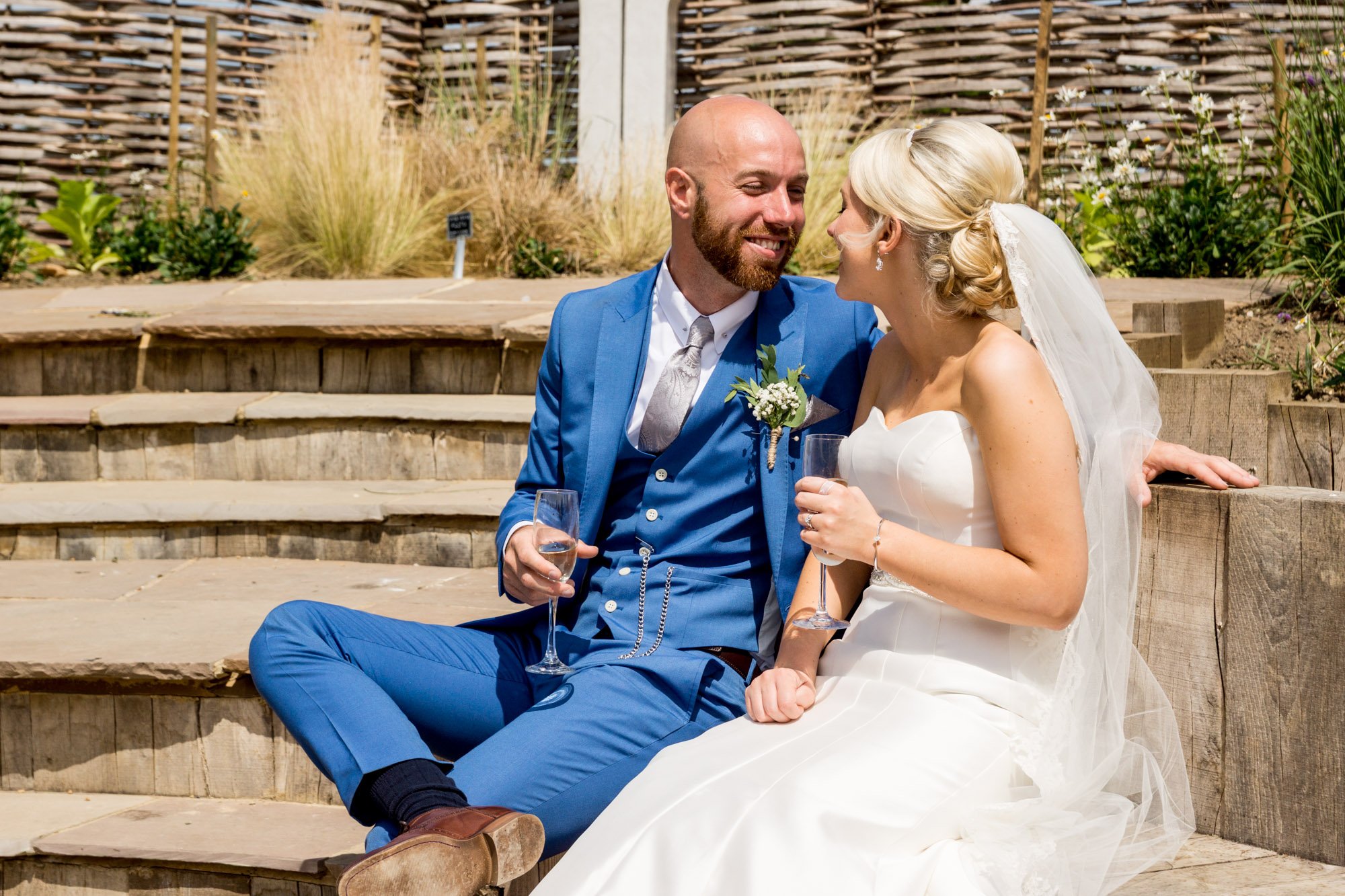 A newlywed couple sitting on outdoor steps, smiling and facing each other, holding glasses of champagne, with a wooden fence and plants in the background.