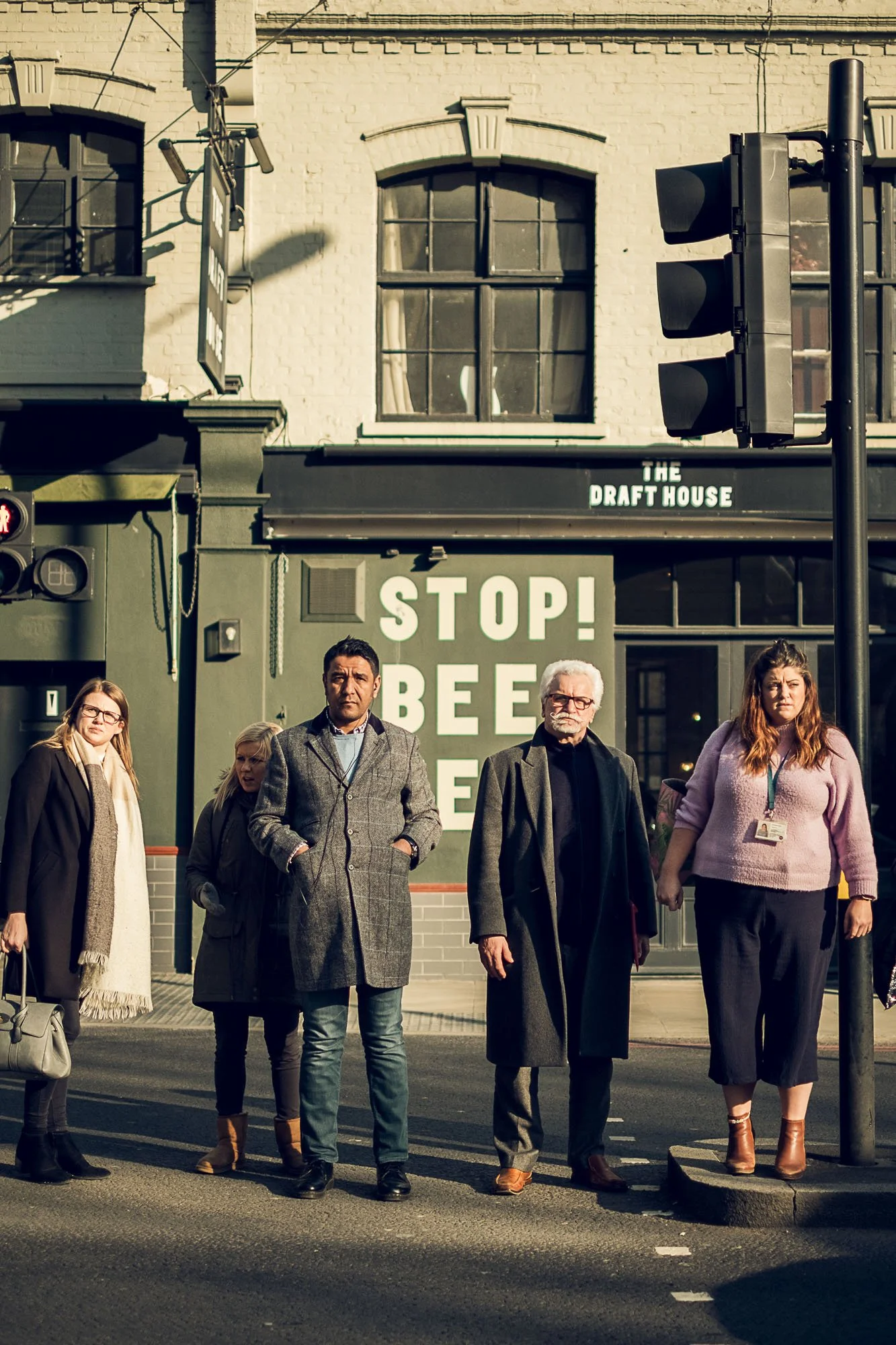 A group of five people standing at a crosswalk during daylight in front of a building with a sign that says "STOP! BEE" and a traffic light with a red pedestrian signal. The scene appears urban.