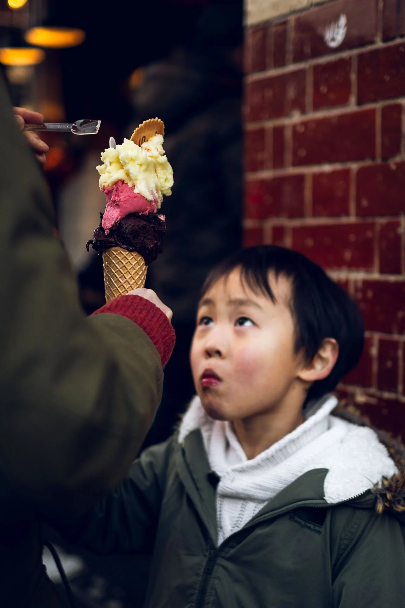 A young boy looking at a three-scoop ice cream cone with chocolate, vanilla, and strawberry flavors, held by someone else, standing against a brick wall.