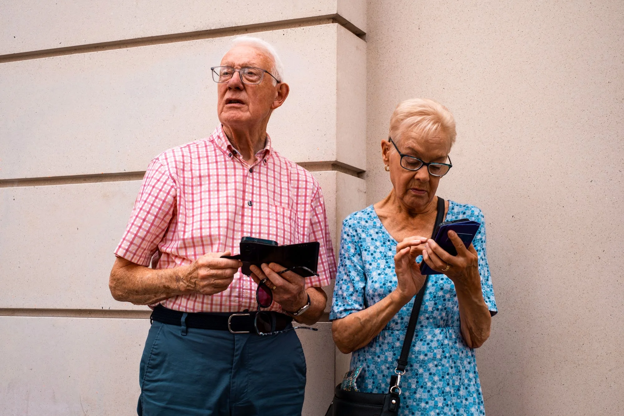 An elderly man and woman standing against a beige wall, both looking at their smartphones.