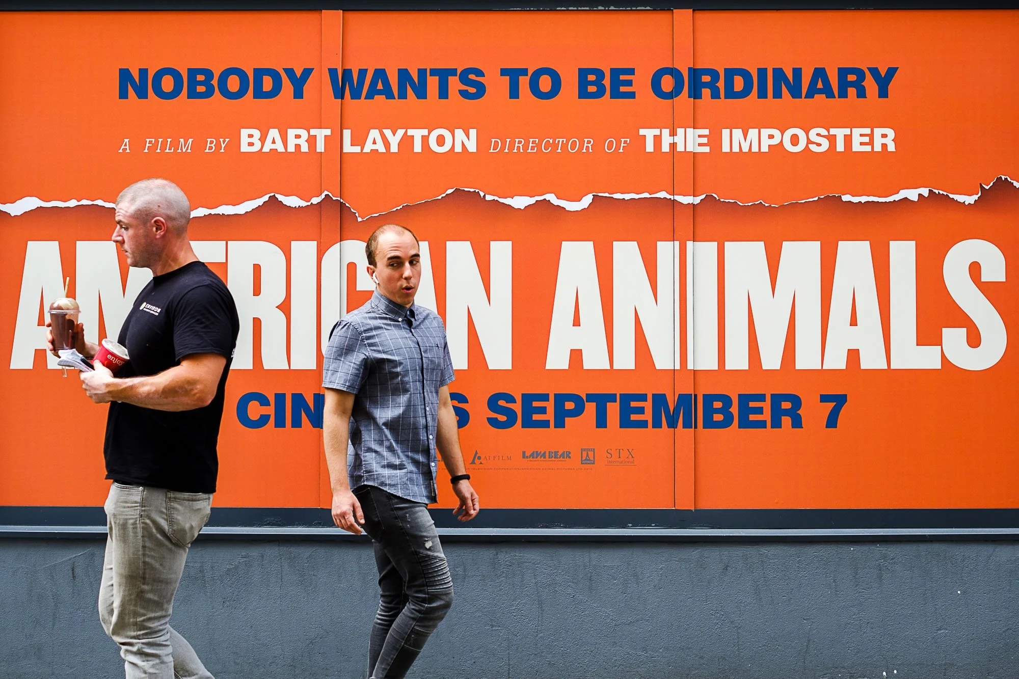 Two men walking past a large orange poster advertising the film 'American Animals' with release date September 7.