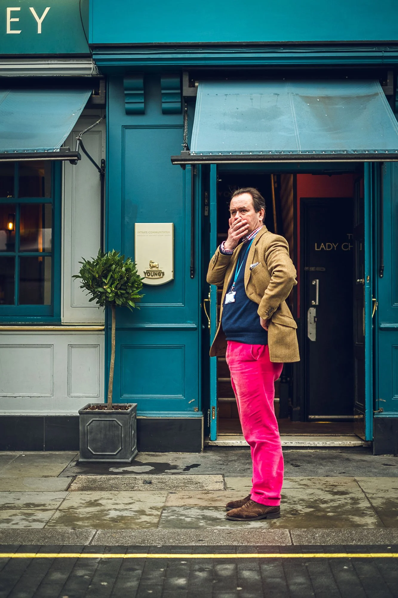 A man standing outside a blue building, wearing a tan blazer, navy sweater, checkered shirt, bright pink pants, and brown shoes, covering his mouth with his hand.