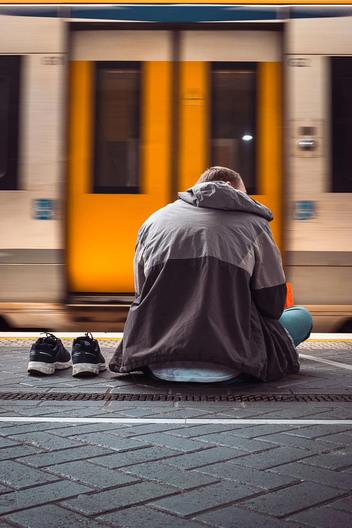 A person sitting on a city sidewalk at night with their back to the camera, shoes removed and placed beside them, as a yellow and blue train passes by in motion.