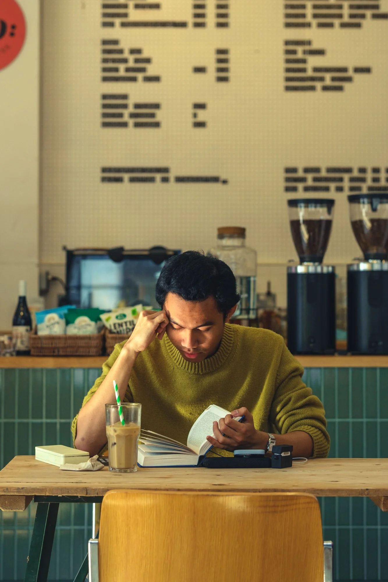 A young man with dark, wavy hair wearing a yellow sweater, sitting at a wooden table in a coffee shop, reading a book while resting his head on his hand. On the table, there is a glass of iced coffee with a green straw, a small white box, and a smart