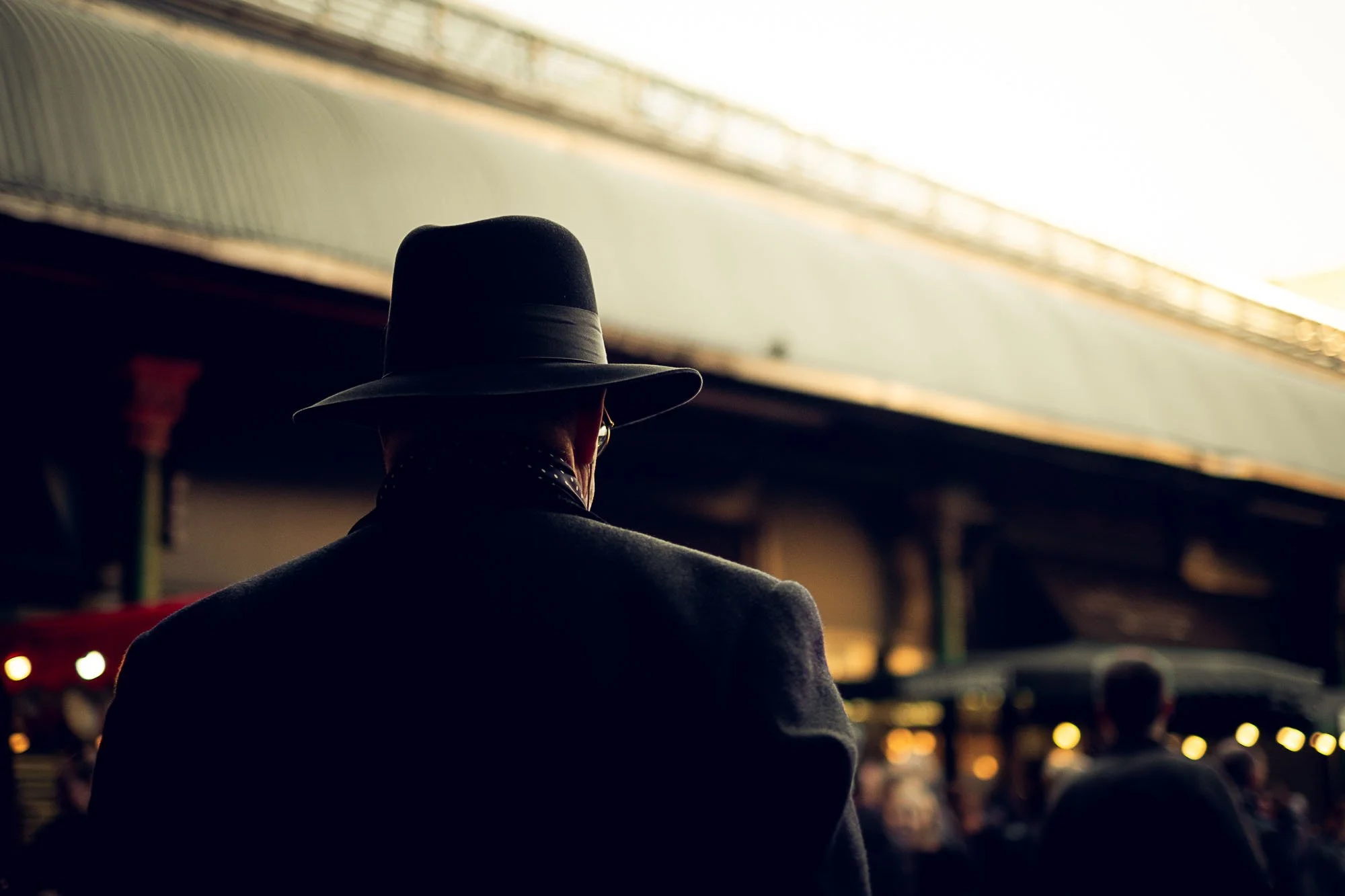 Back of a man wearing a black fedora hat and dark coat, standing in an outdoor market at sunset.