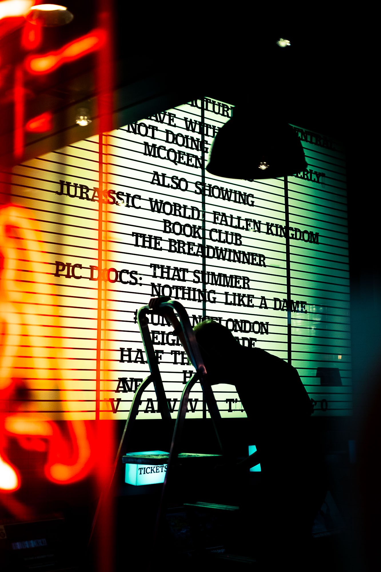 A person arranging a brightly lit movie theatre marquee with movie titles and showtimes, and a ladder with a small illuminated sign that says "TICKETS."