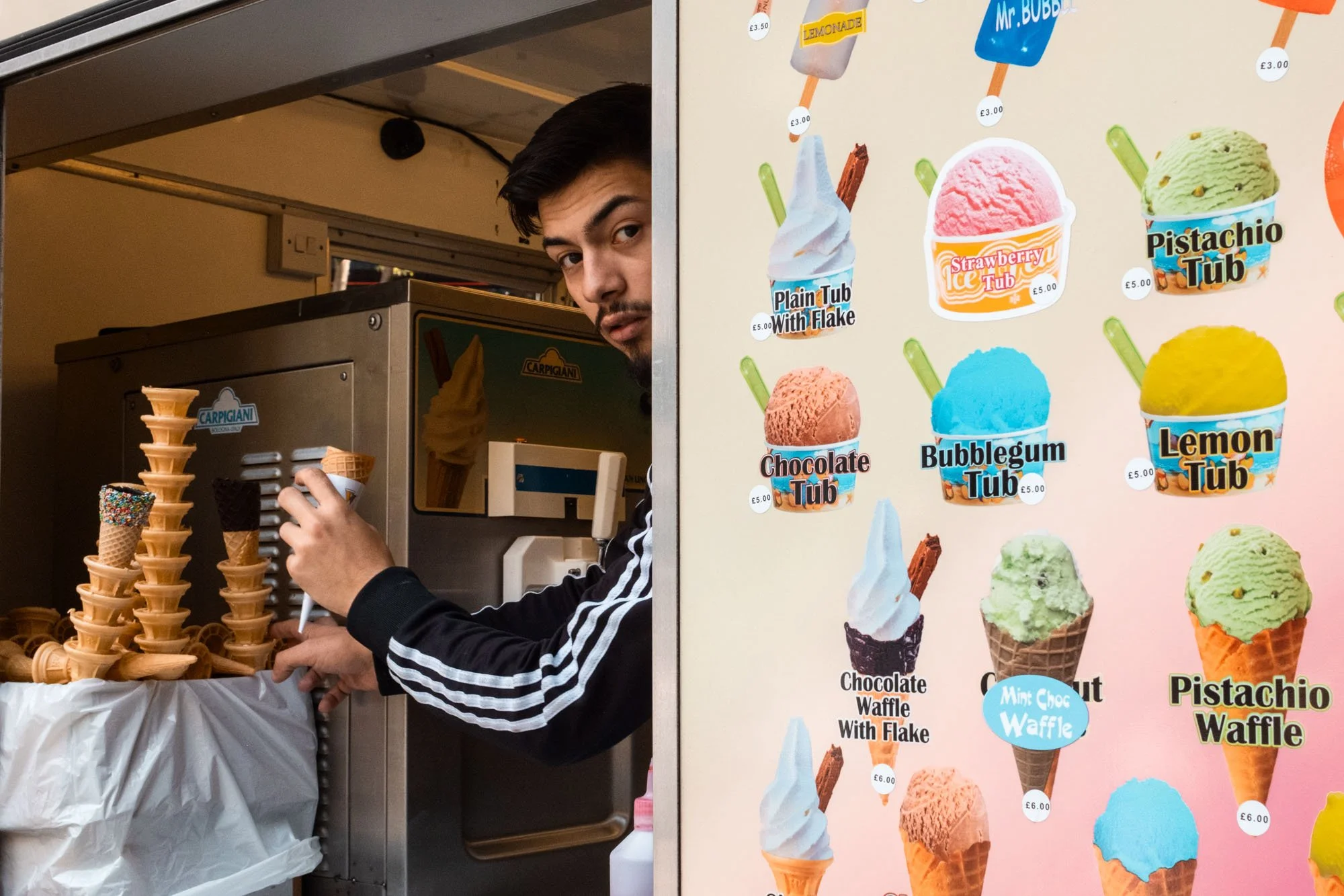 A young man with dark hair and a beard serving ice cream at a stand. There is a rack of cone ice cream on his left and a colorful menu on his right displaying various ice cream flavors and prices.