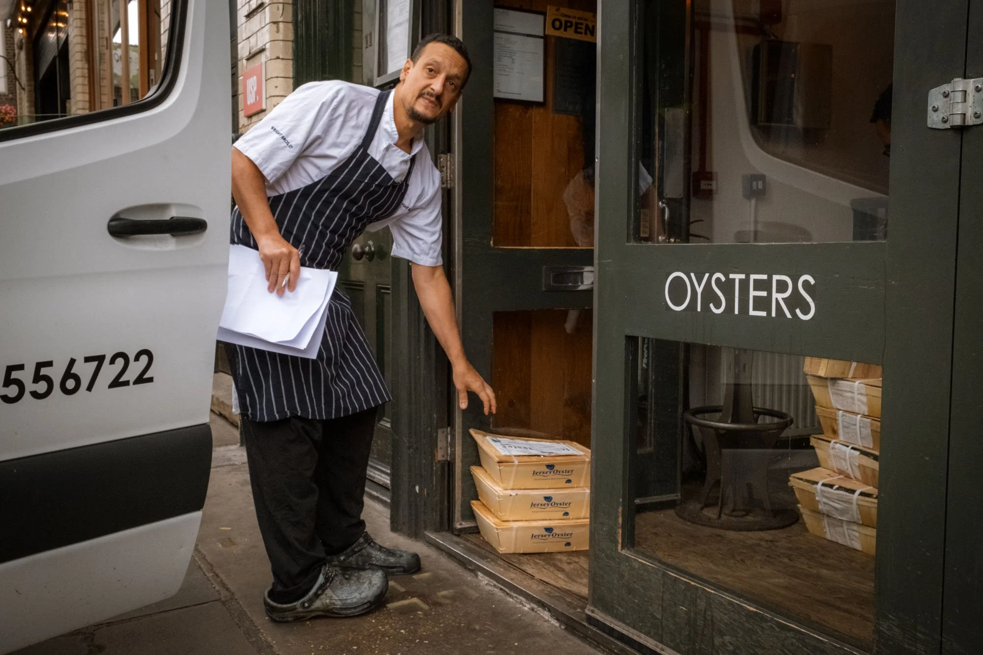 A man in a striped apron delivers boxes of oysters from a van to a shop labeled 'Oysters'.