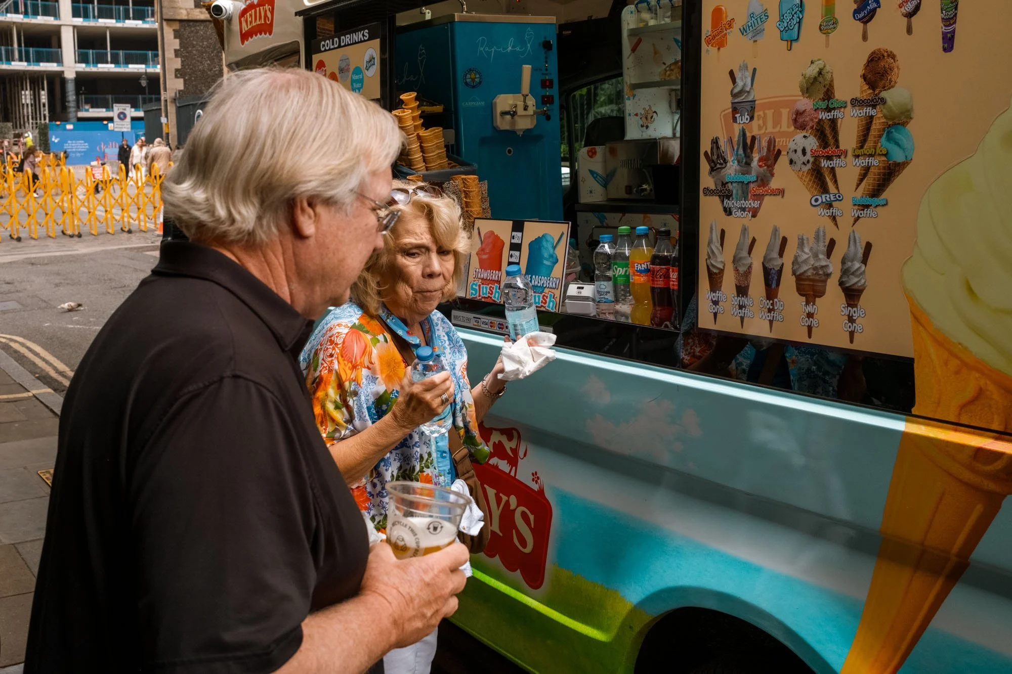 Two older adults, a man and a woman, standing in front of an ice cream truck. The woman holds a water bottle and a napkin, and the man holds a beer. The ice cream truck displays various ice cream cones and milkshake options on a colorful menu.