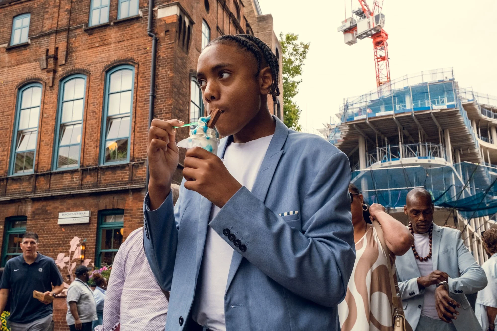 A young man with braided hair in a grey blazer eats ice cream with a chocolate flake at an outdoor event, with a construction site on a tall building in the background and several people around.