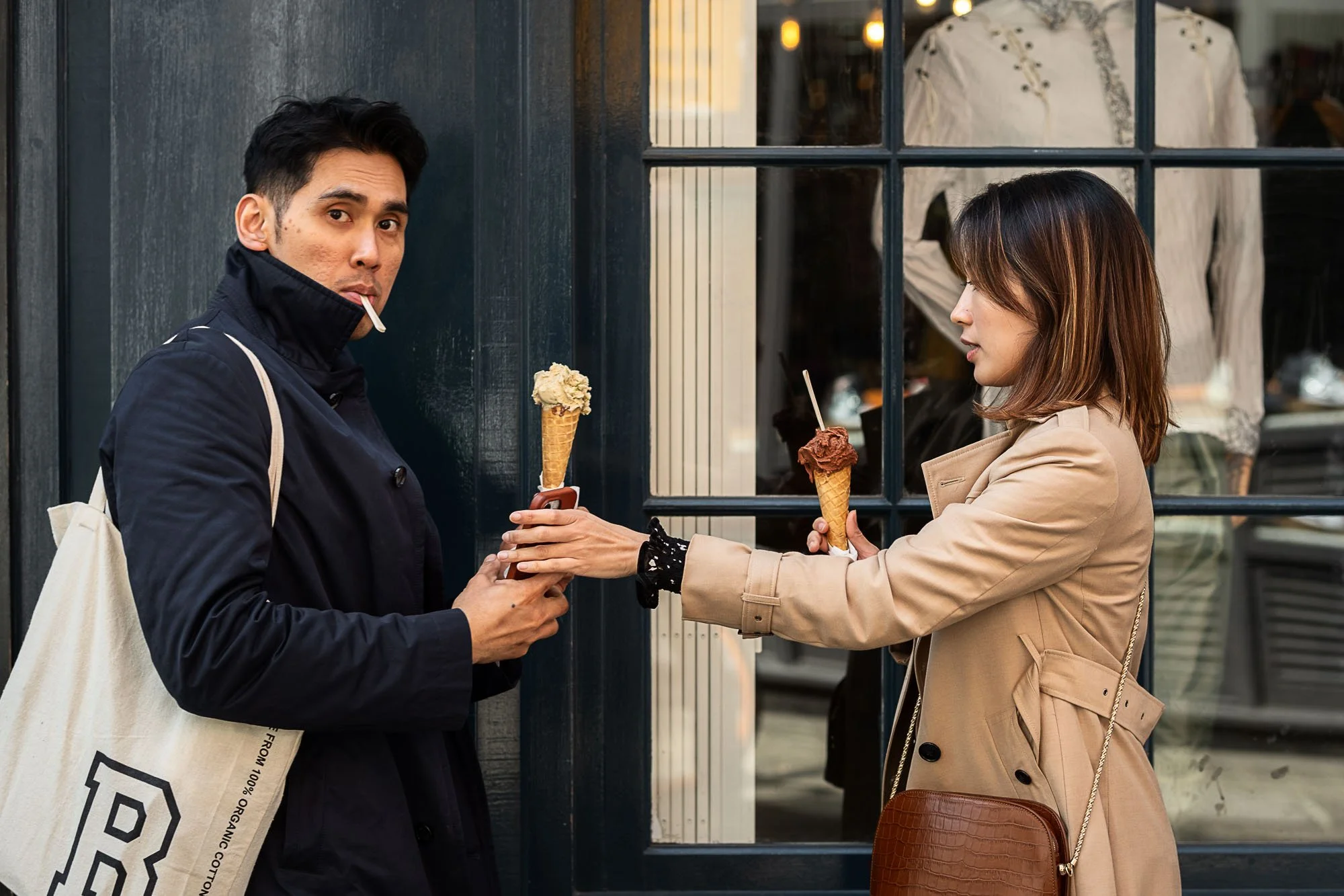 A man in a dark jacket and a woman in a beige trench coat share ice cream cones outside a shop, with the man looking at the camera and the woman in profile.