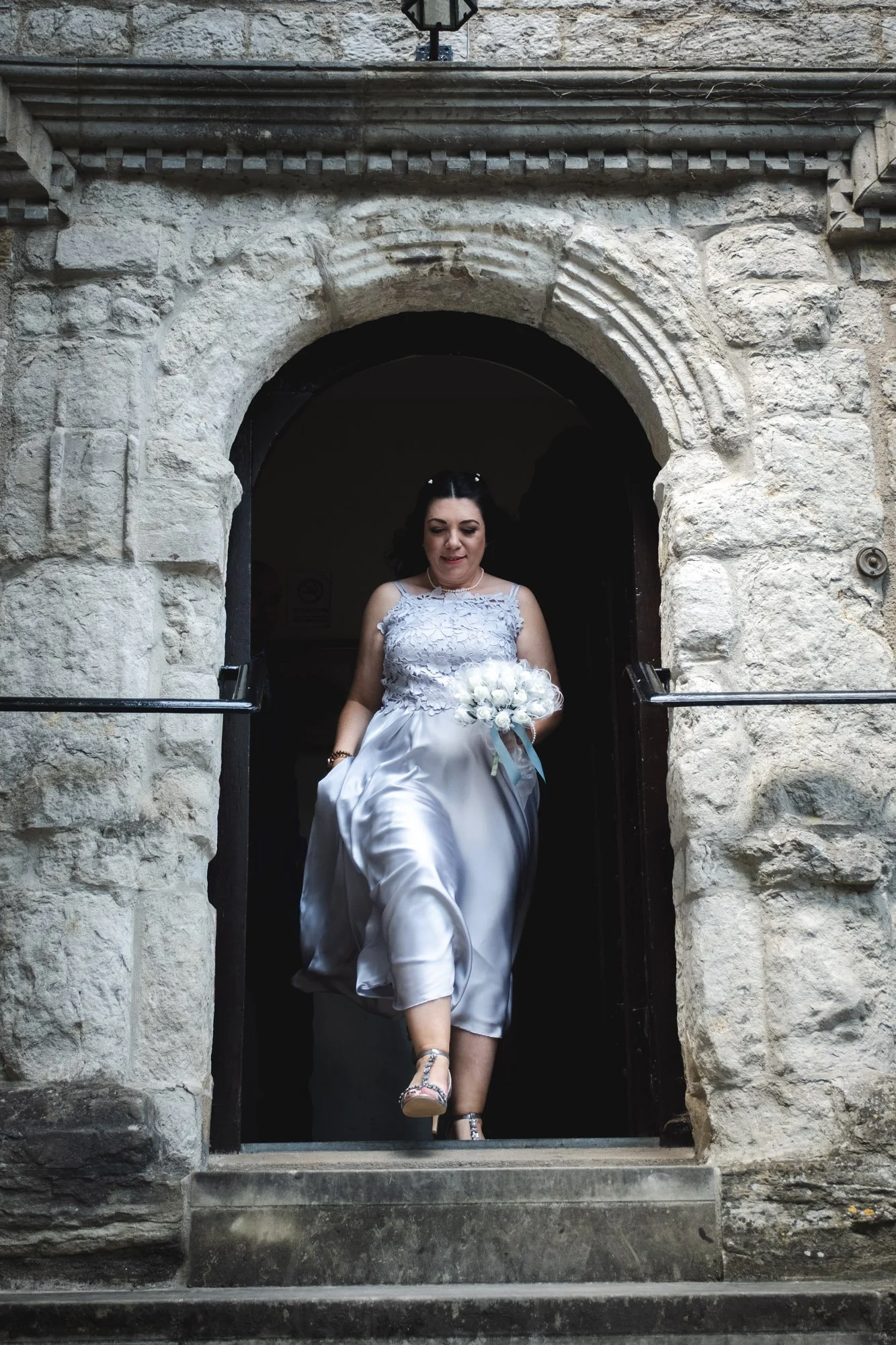 A woman in a white dress walking out of a stone archway holding a bouquet of white flowers.