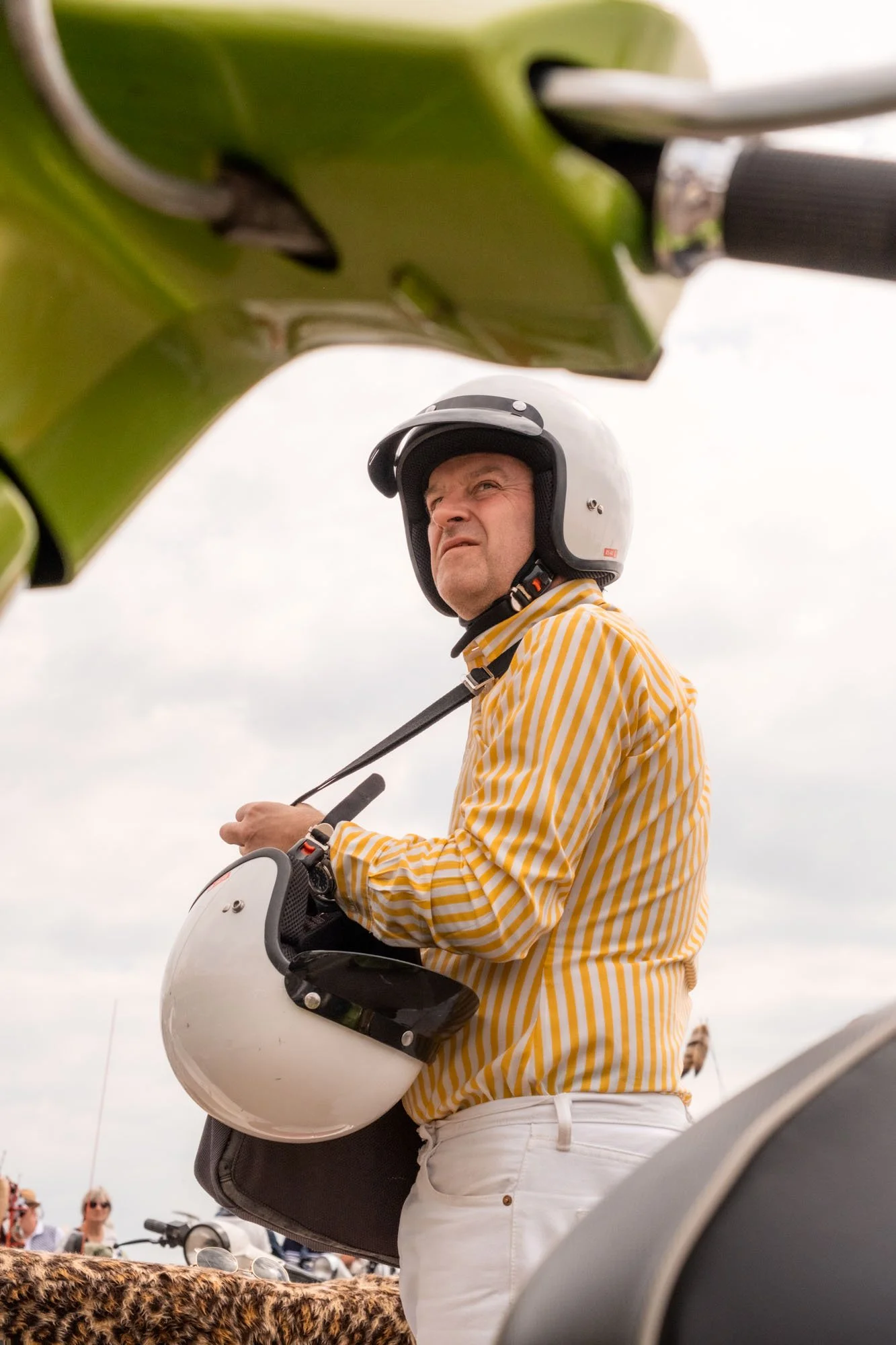 A man wearing a yellow and white striped shirt, white pants, and a white safety helmet, holds a white helmet and looks upward, with a green motorcycle in the foreground and people in the background.