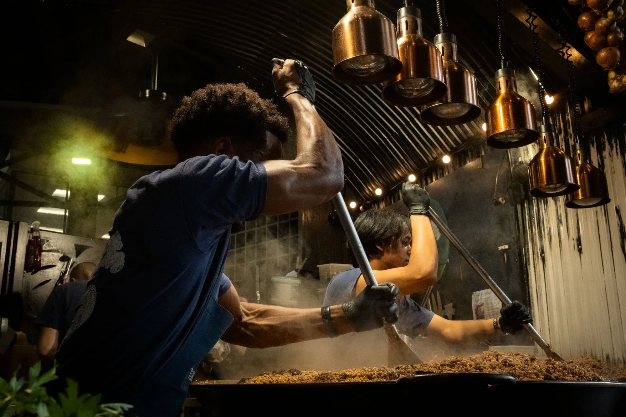Two chefs are cooking a large dish of food in a commercial kitchen, stirring the food in a pan. The kitchen has dark lighting with hanging copper pots and a corrugated black ceiling.