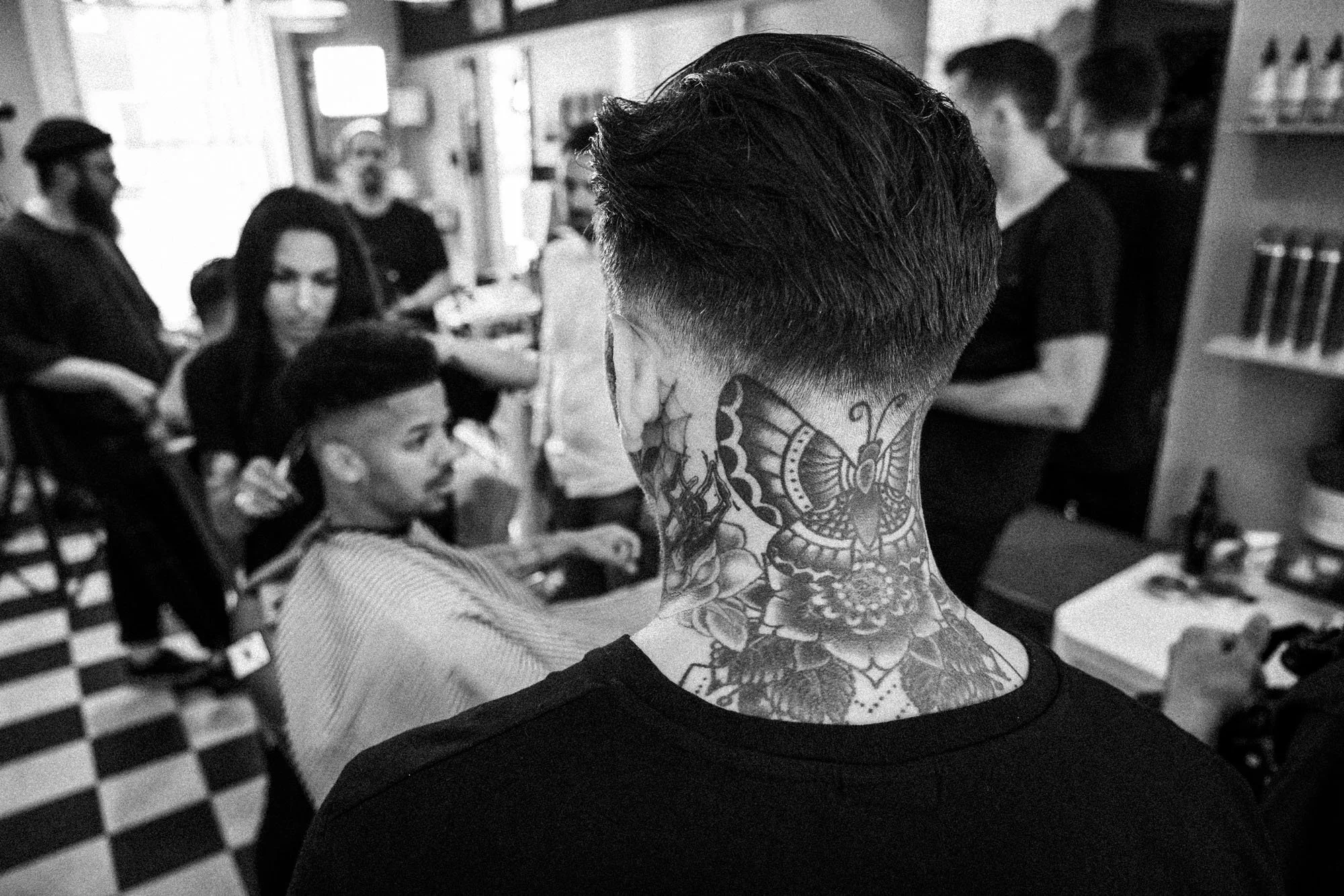 A person with a detailed butterfly tattoo on the neck, facing away in a barbershop with people, including a boy with a striped cape, in the background.