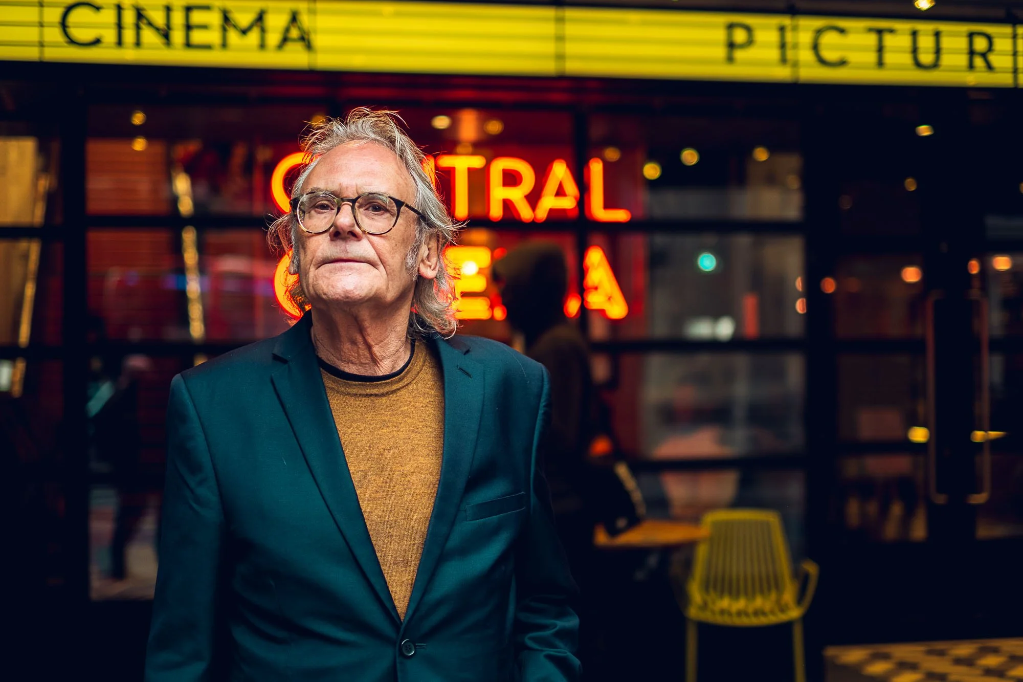 Older man with glasses and gray hair standing outside a cinema at night, illuminated neon sign and colorful lights in the background.
