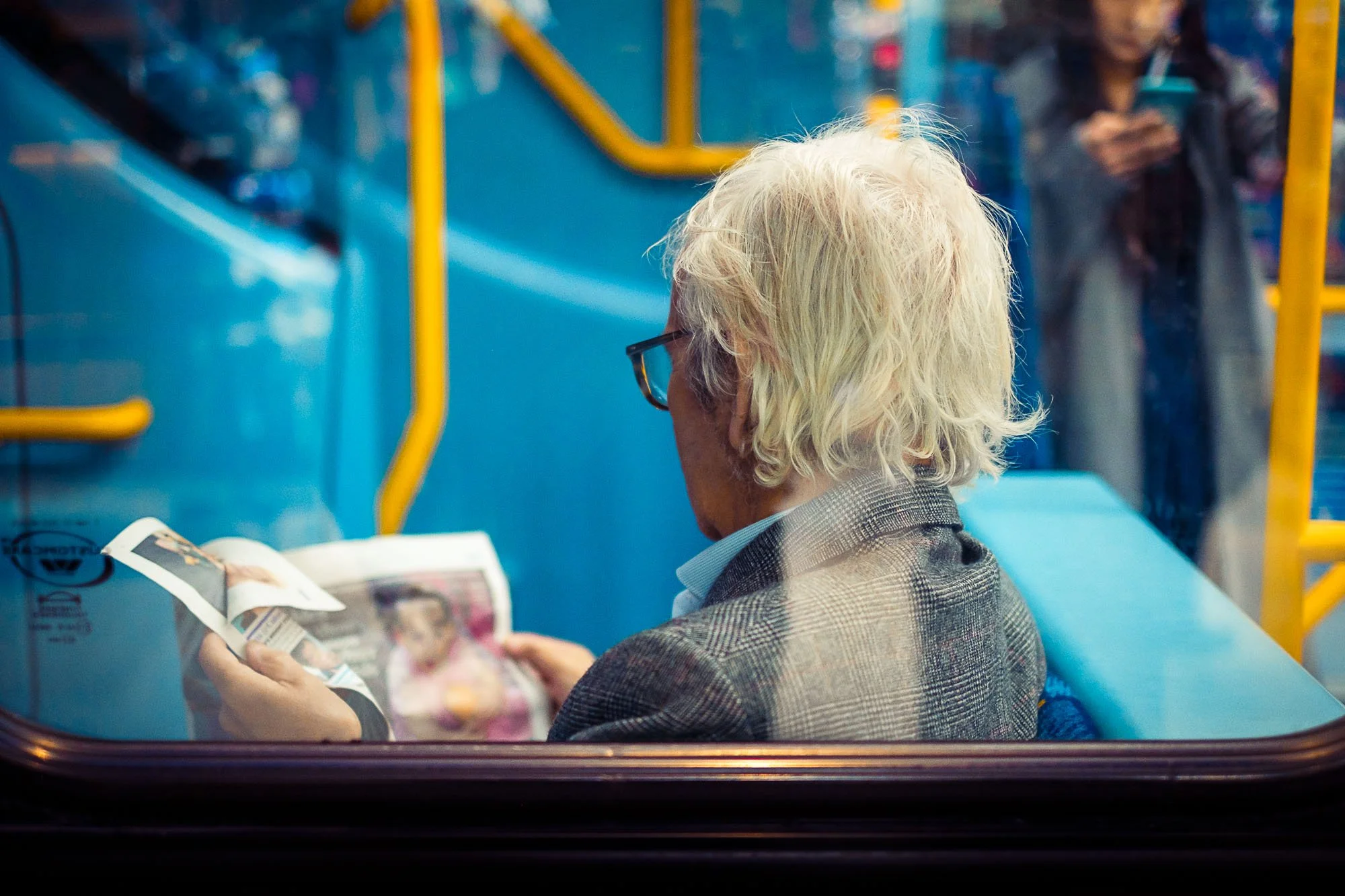 An elderly man with white hair and glasses sitting on a bus, reading a magazine. A woman in the background is looking at her phone.