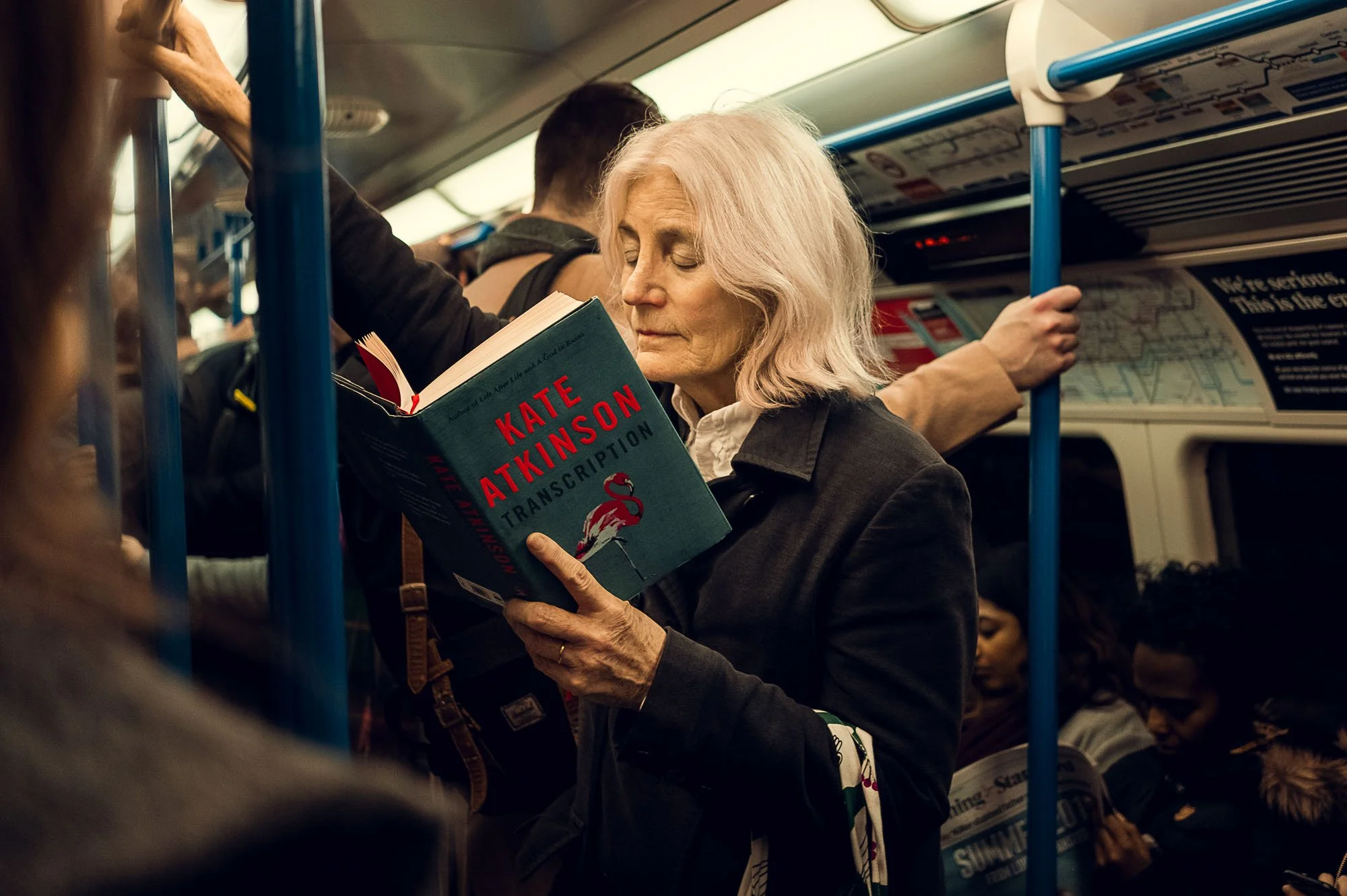 An elderly woman with light hair is reading a book titled 'Transcription' while standing in a crowded subway train. She appears focused with her eyes closed, holding onto a vertical blue pole with one hand. Other passengers are visible, some standing