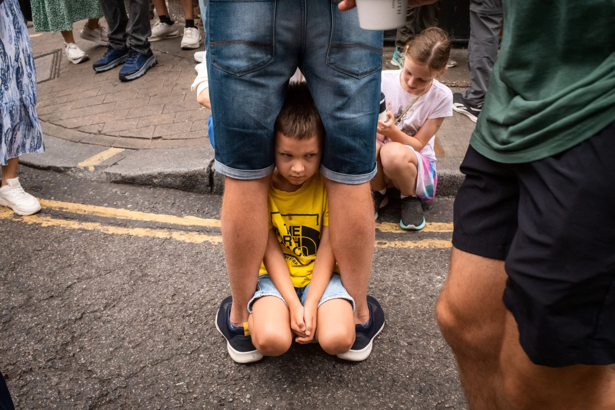A young boy sitting on the ground with a serious expression under an adult's legs, surrounded by other people in an outdoor urban setting.