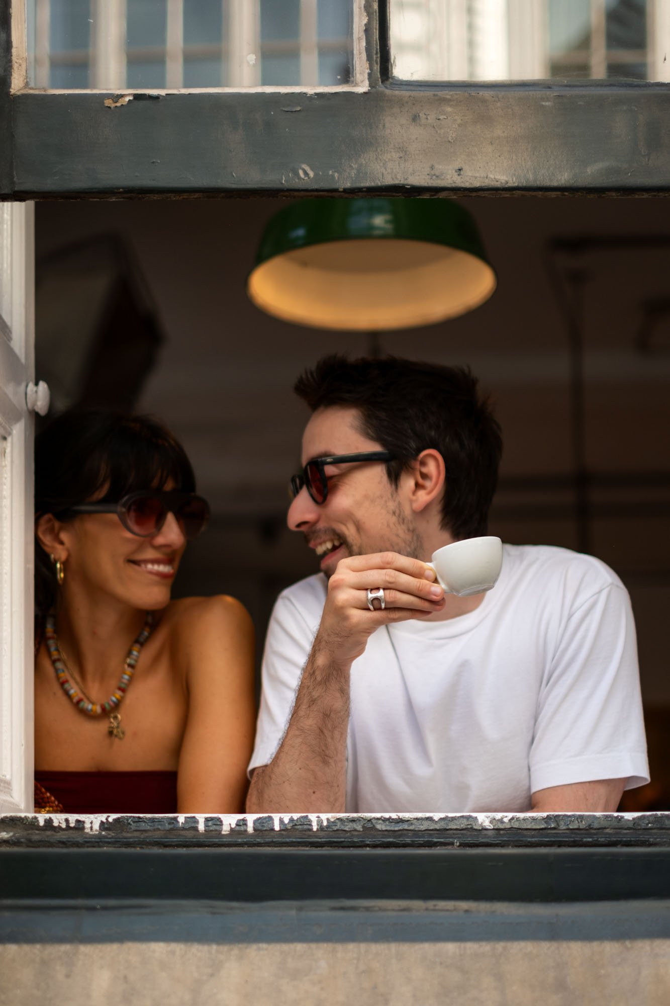 A man and woman are smiling at each other through a window, with the man holding a white teacup, in a cozy indoor setting.