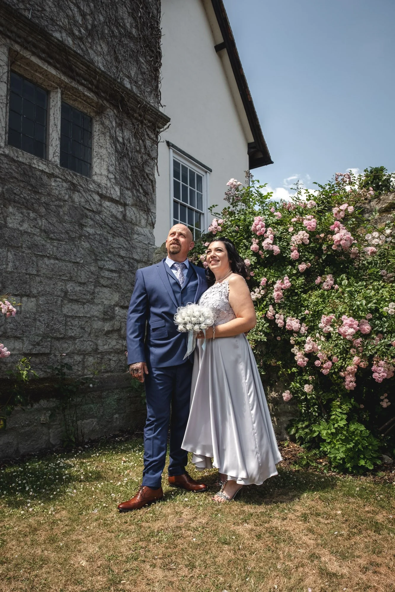 A bride and groom standing outdoors in front of blooming pink roses and a stone wall, smiling and dressed in wedding attire, with a white house and blue sky in the background.