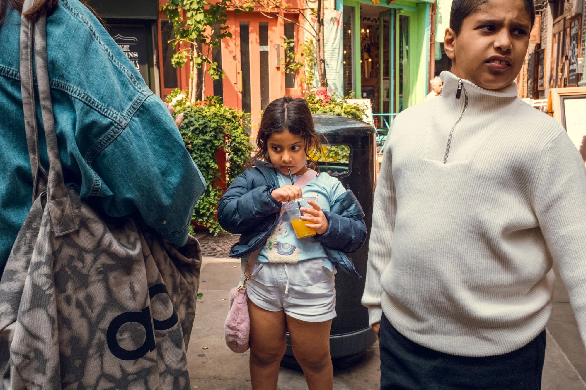 A young girl drinking orange juice with a straw while surrounded by two boys on a city street with colorful buildings in the background.