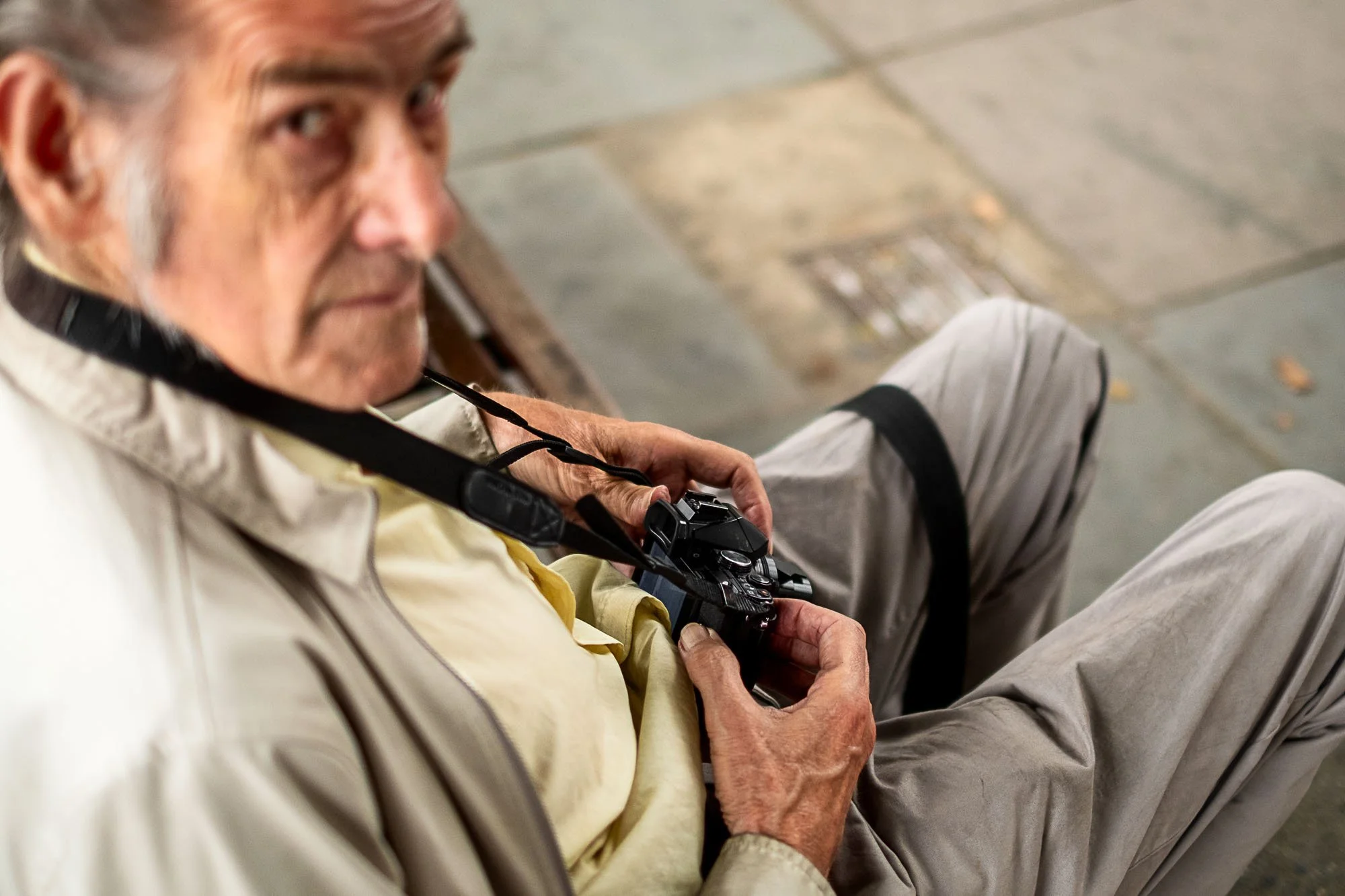 An elderly man sits on a sidewalk holding a camera with a strap around his neck, looking upwards.