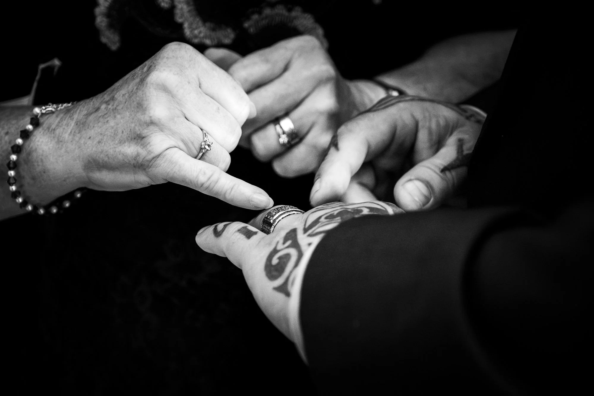 Close-up of two people holding hands, one with tattooed arm and ring, the other with beaded bracelet and rings, in black and white