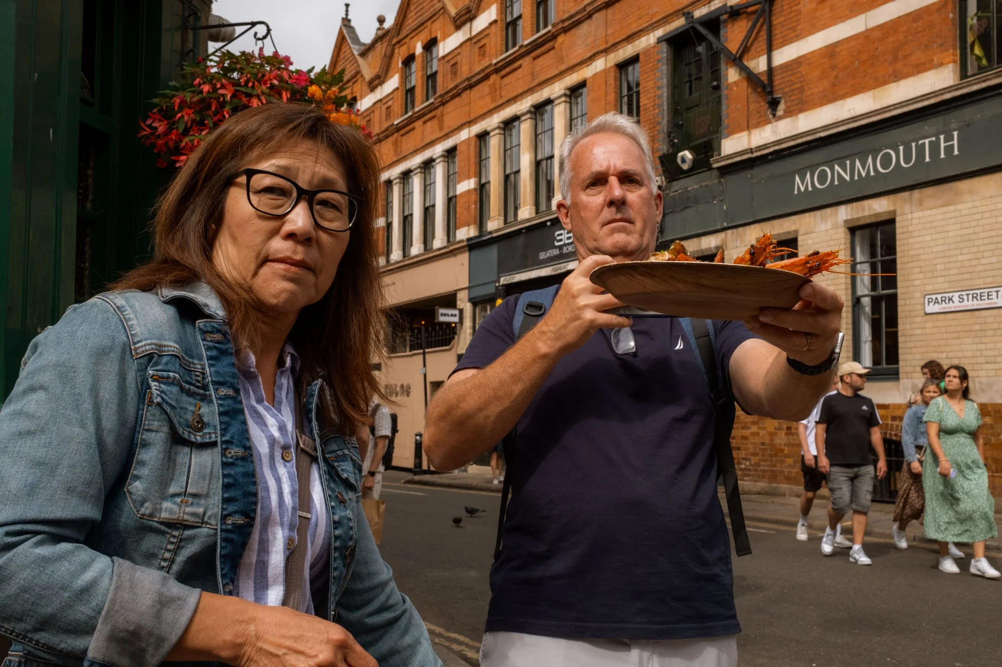 A man holding a plate of lobster and a woman standing next to him on a city street. Several pedestrians are in the background, along with brick buildings.