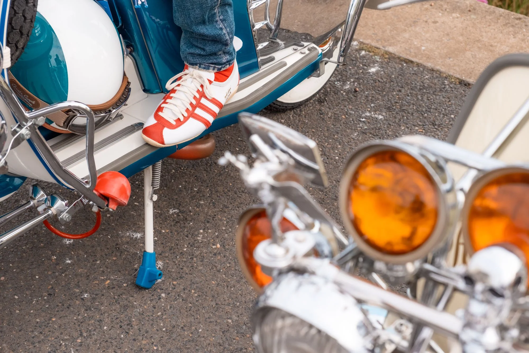 A close-up of the front of a vintage motorcycle with a chrome headlight and orange turn signals, and a person's foot wearing a white and red sneaker resting on the footrest of an older blue and white motorcycle or scooter, parked on an asphalt surfac