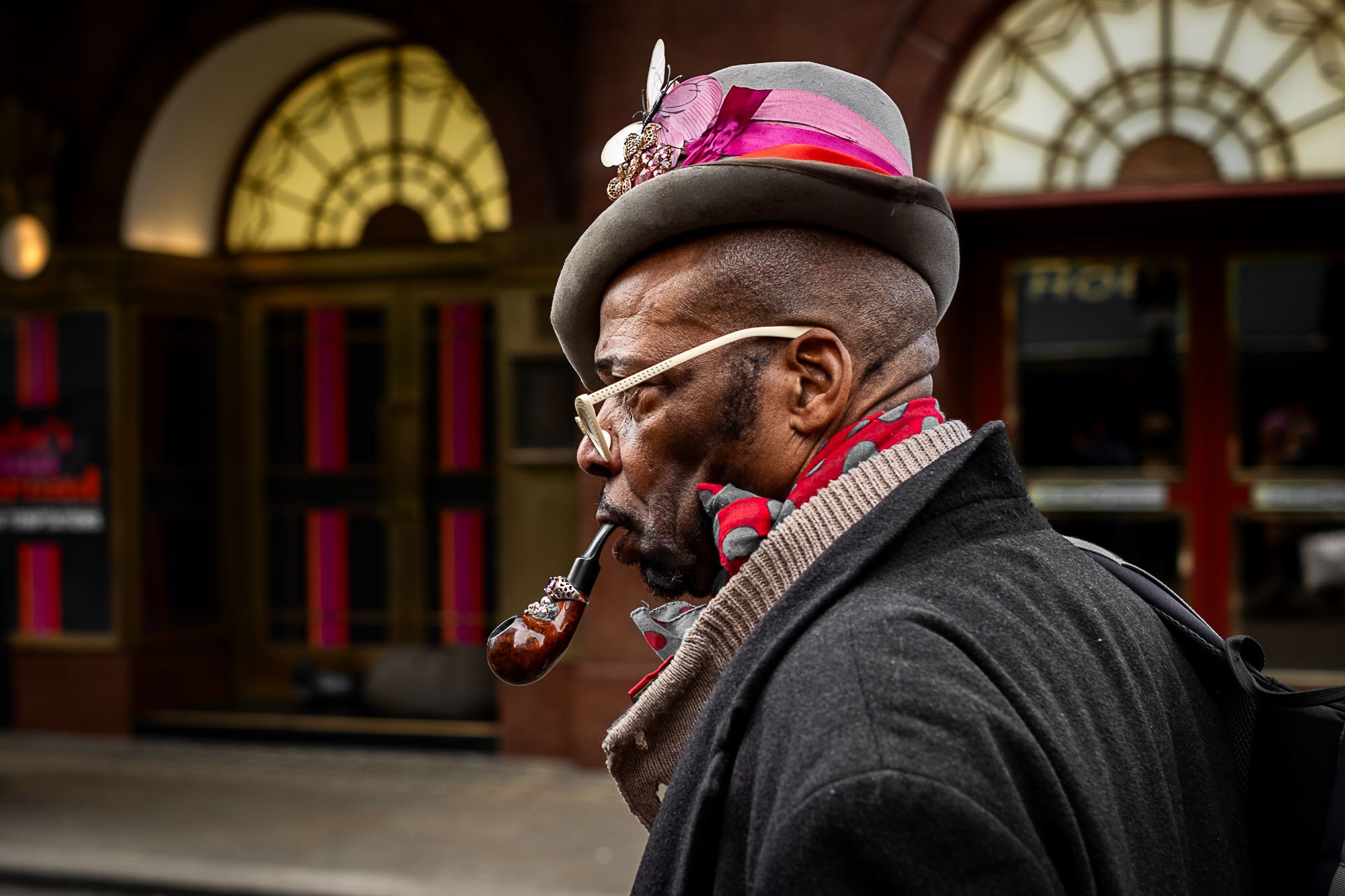 A man wearing a gray hat with pink and purple ribbon accents, cream-colored glasses, and a red and gray scarf, smoking a pipe. He is dressed in a dark coat and appears to be in an indoor setting with large arched windows and warm lighting.