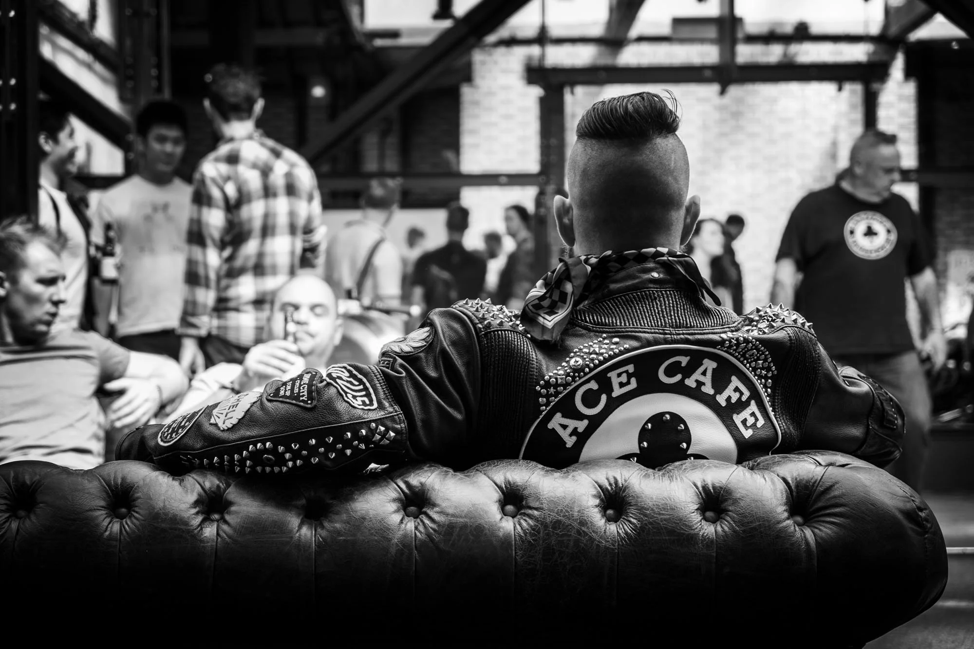 A person with a mohawk hairstyle and a leather jacket with spikes and patches, sitting on a tufted leather sofa in a social setting with other people in the background.