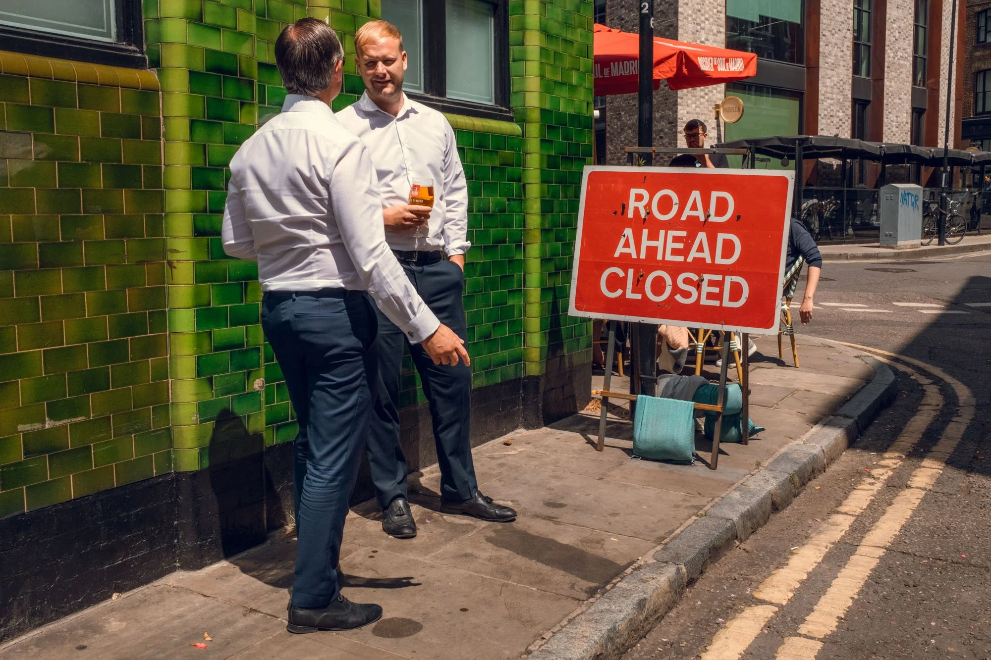 Two men in business attire, one holding a drink, are conversing on a city sidewalk in front of a green tiled building, with a red sign next to them that says 'ROAD AHEAD CLOSED.'