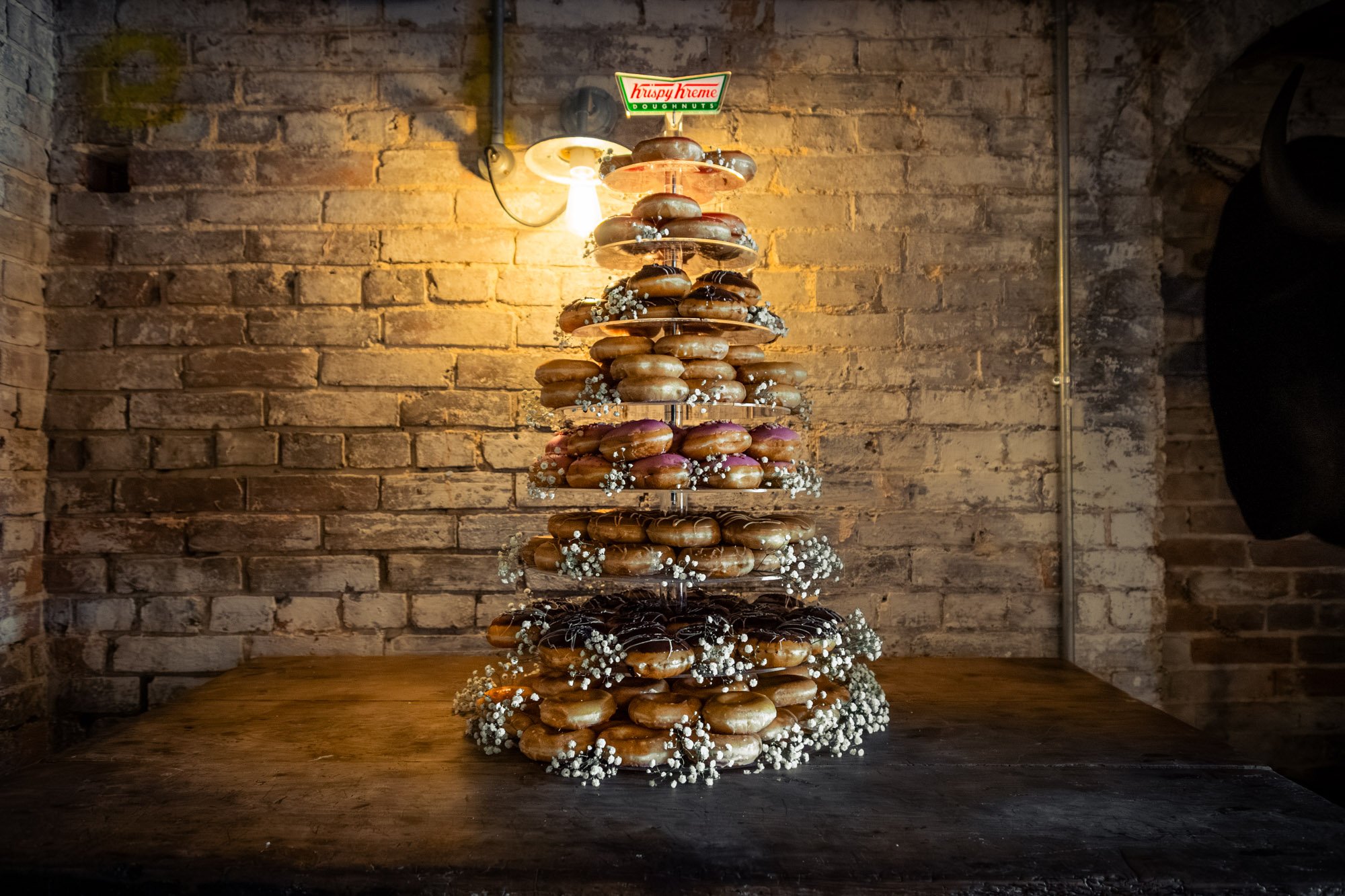A multi-tiered donut display with various donuts, decorated with white flowers, set against a rustic brick wall in a bakery or cafe.