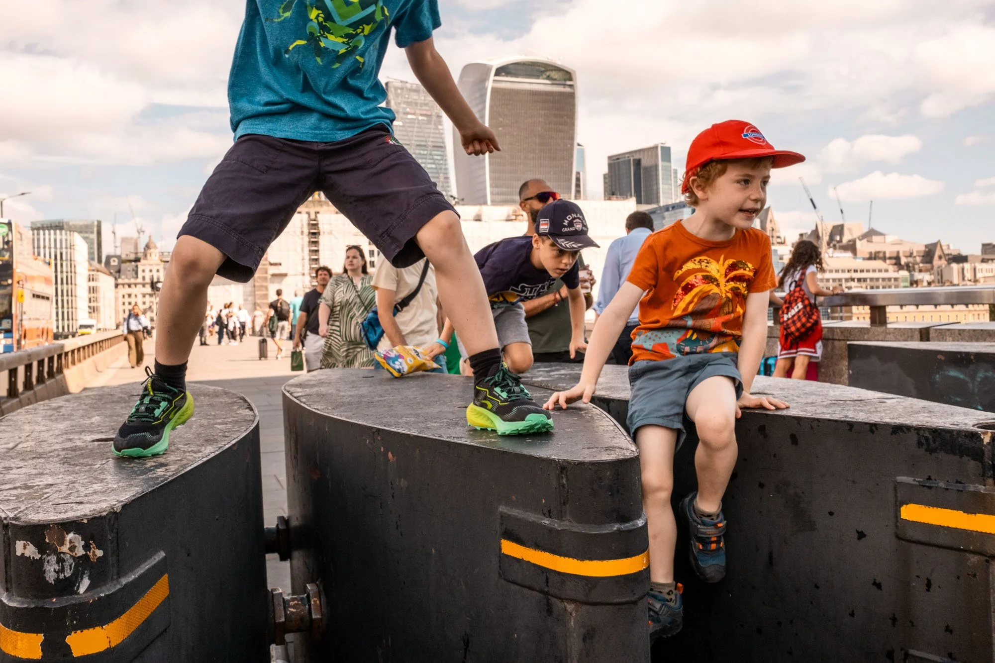 Children playing and climbing on large black concrete blocks outdoors in an urban area with buildings and people in the background.