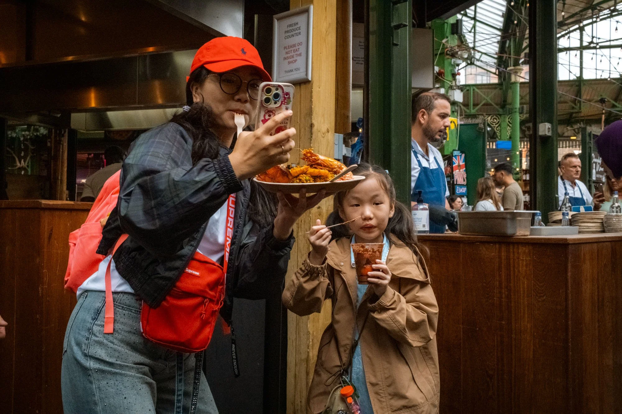 Woman and young girl eating in a busy indoor market or food hall. The woman is taking a photo with her phone, holding a plate of food, while the girl is holding a cup and eating with chopsticks. Market stalls and other people are visible in the backg